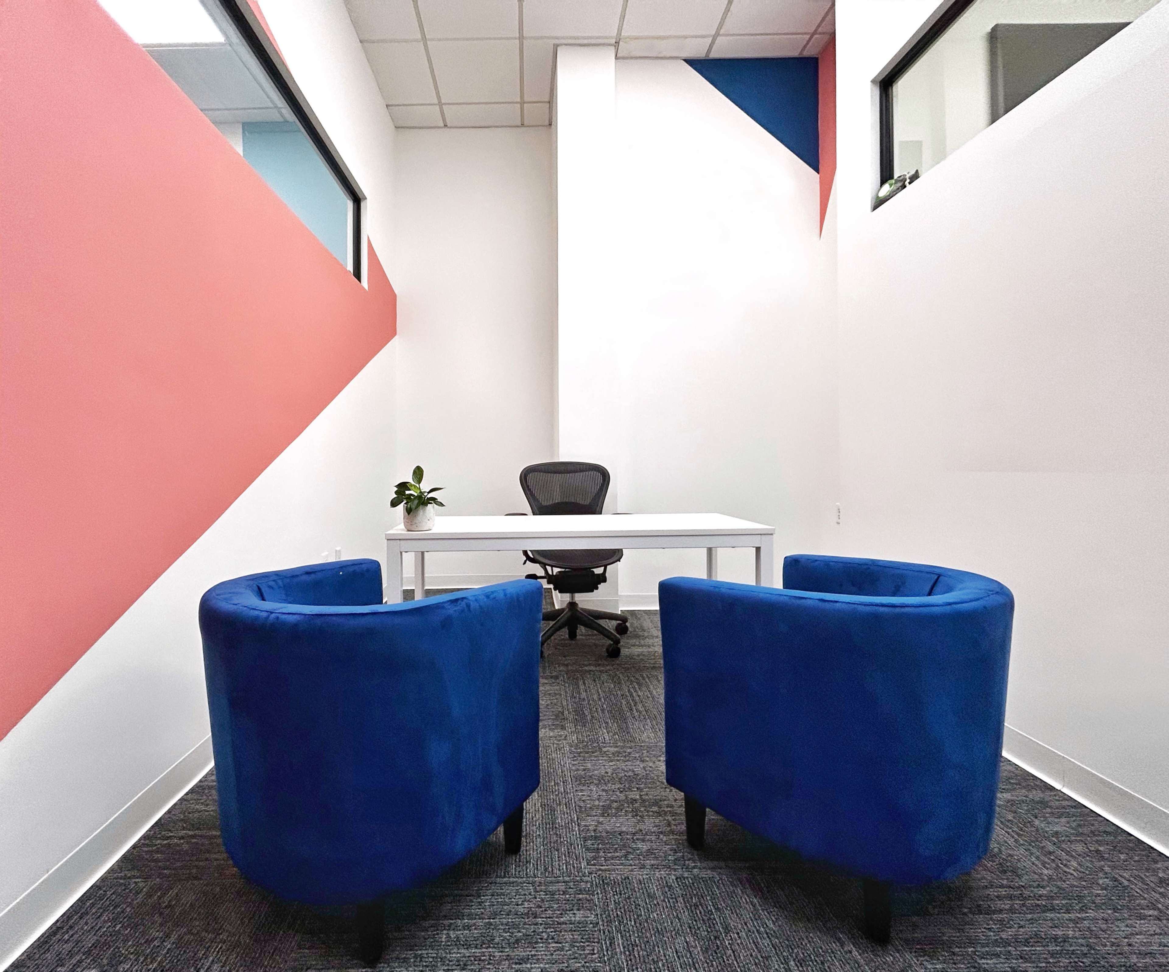 A minimalist office space with a white desk and a black ergonomic chair, flanked by two blue chairs, set against a backdrop of colorful diagonal wall patterns.