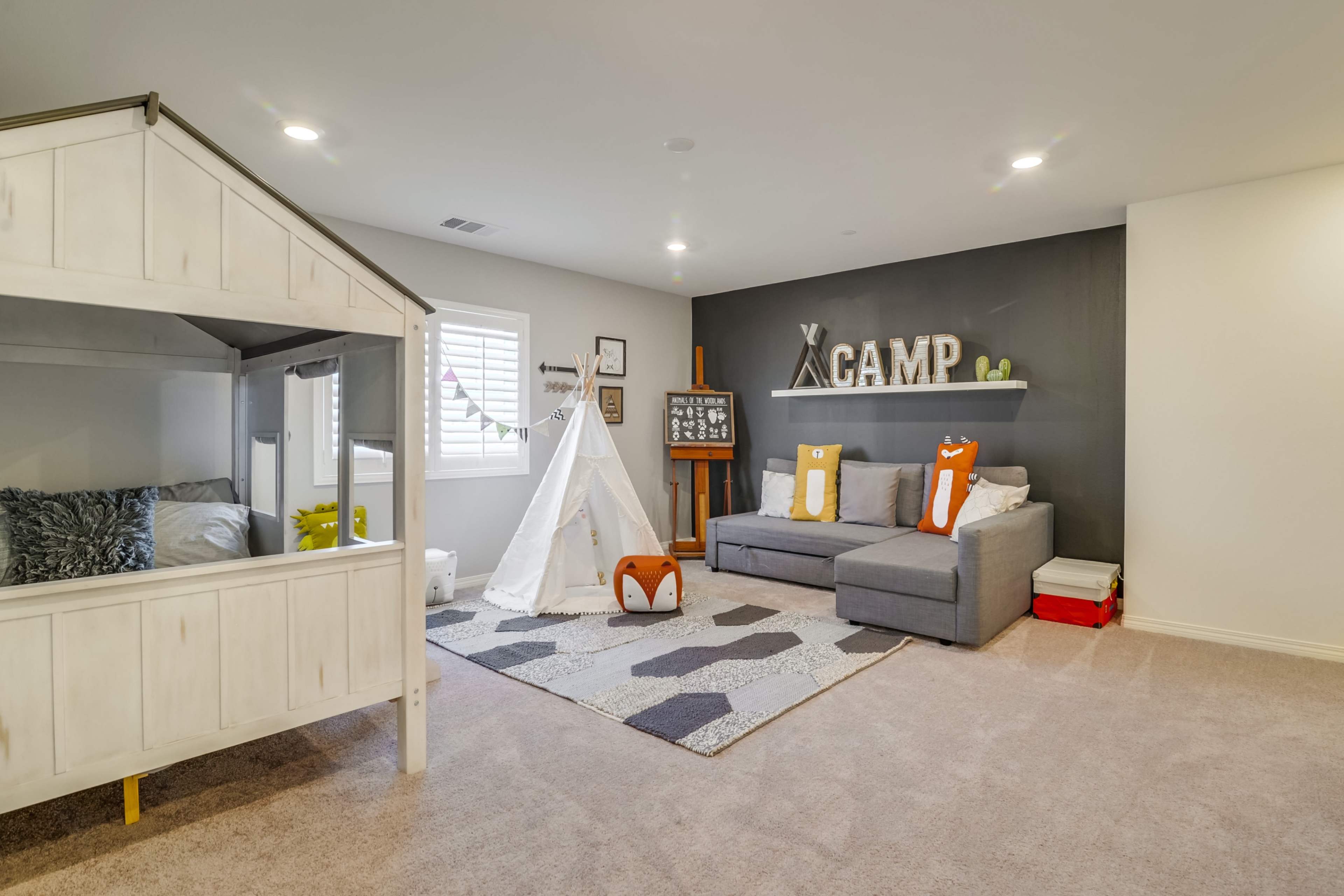 A cozy playroom featuring a house-shaped play structure, a white teepee, a gray sectional sofa, and a decorative "CAMP" sign on the wall.