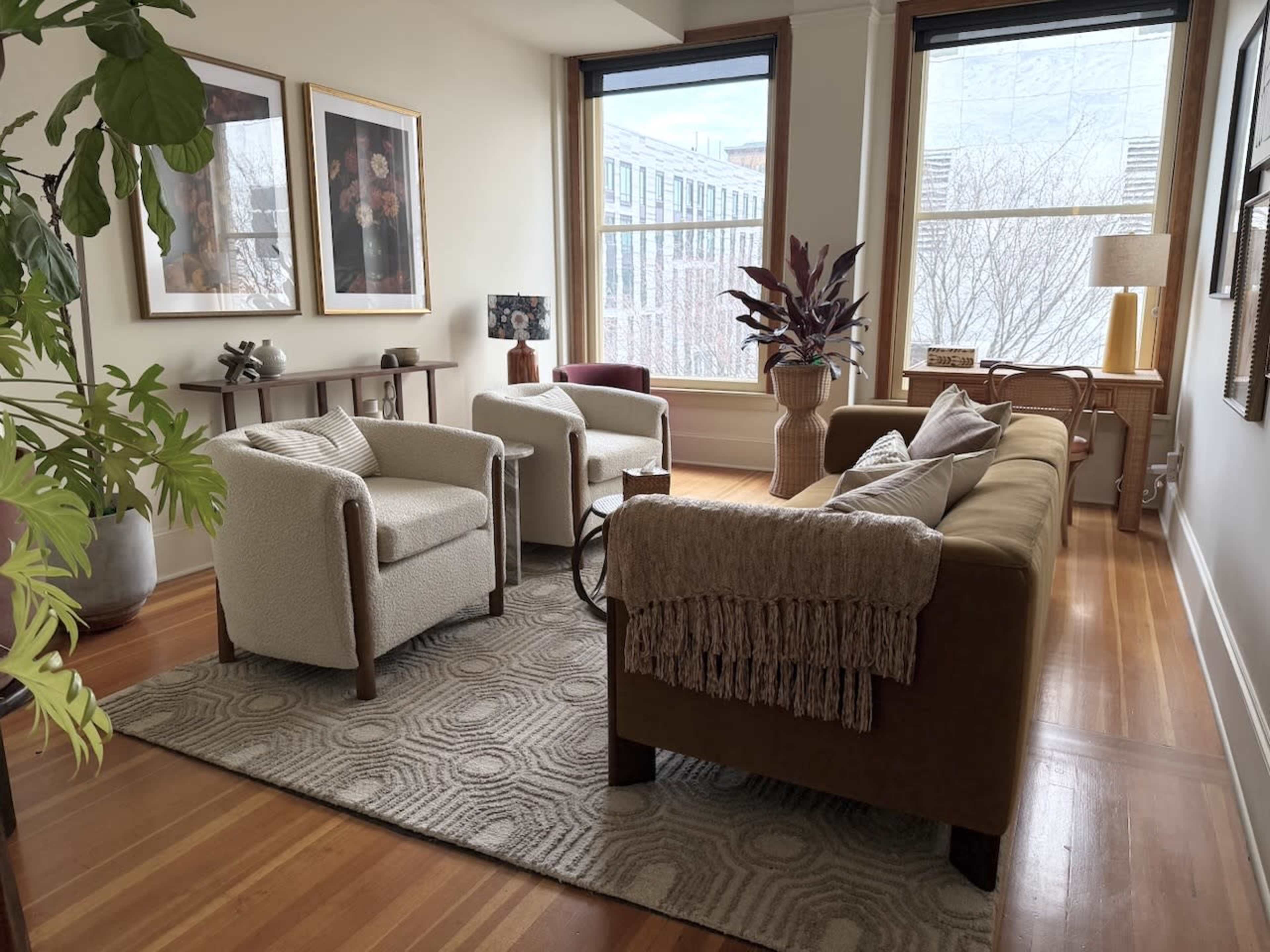 The image shows a cozy interior living room with two armchairs, a sofa, decorative art on the walls, and large windows letting in natural light.