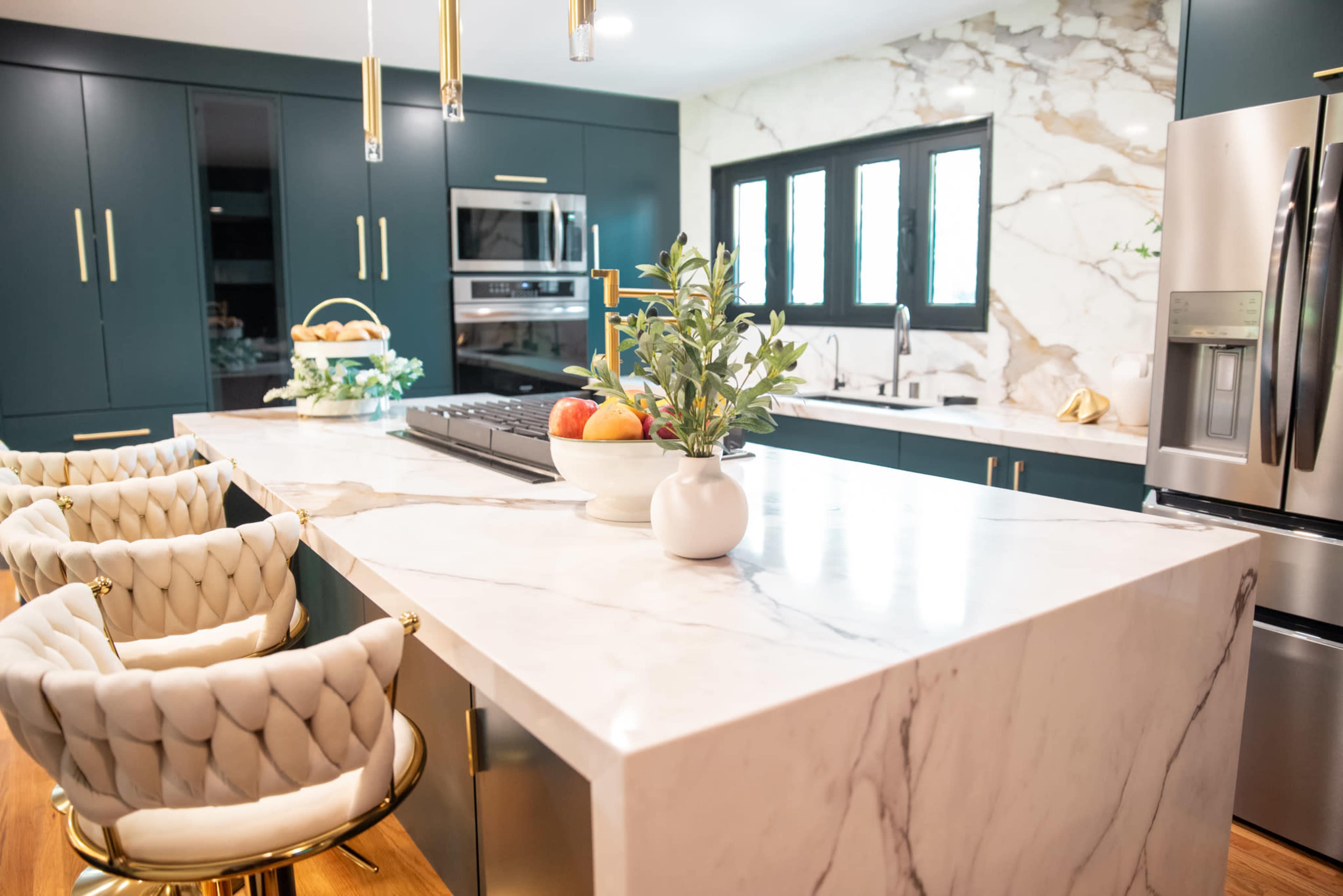 A modern kitchen features a large marble island with gold-accented bar stools, dark cabinetry, and marble wall panels.