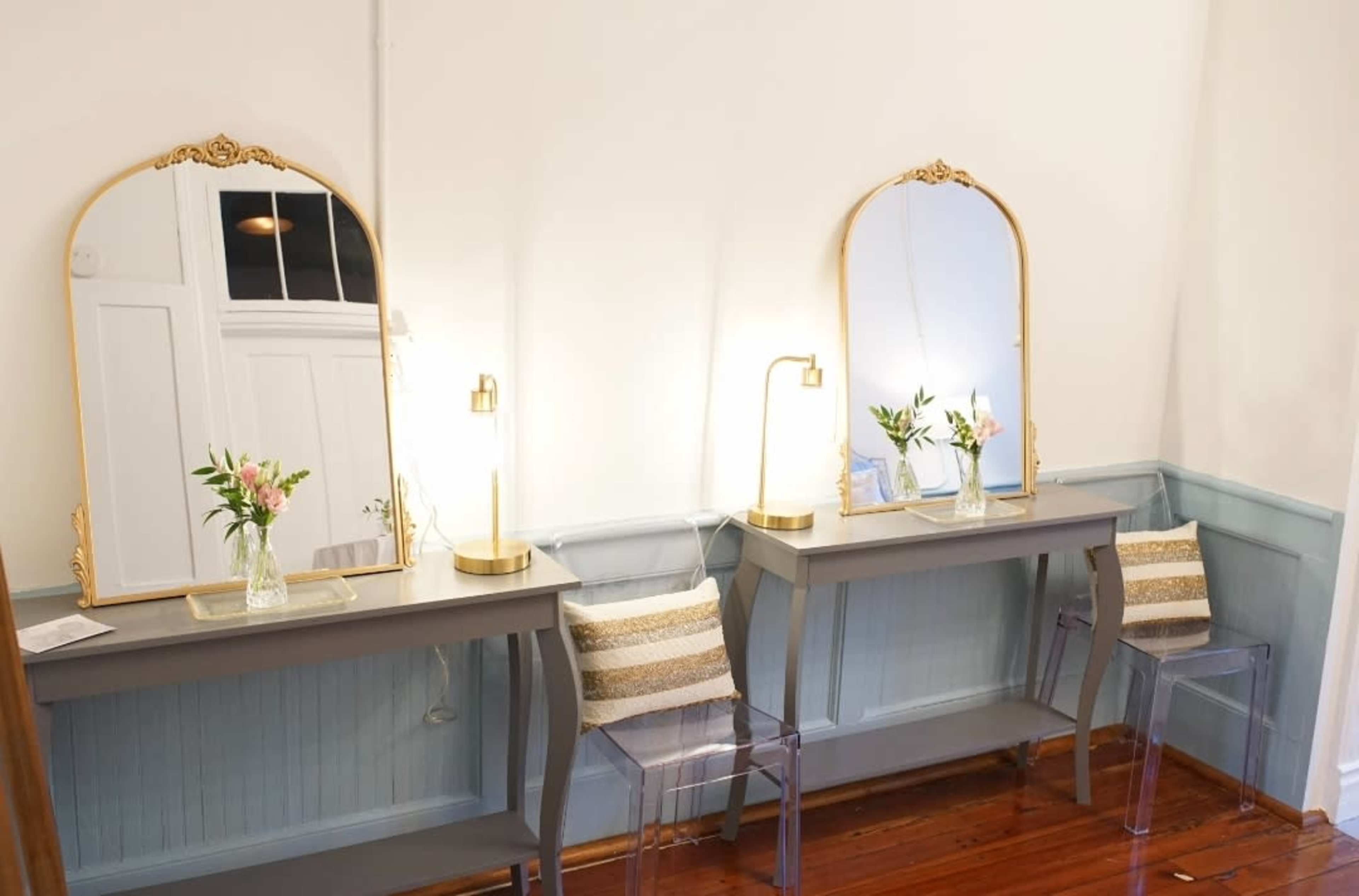Two gray vanities with gold-framed mirrors and table lamps are positioned against a light-colored wall, each accompanied by a clear acrylic chair.