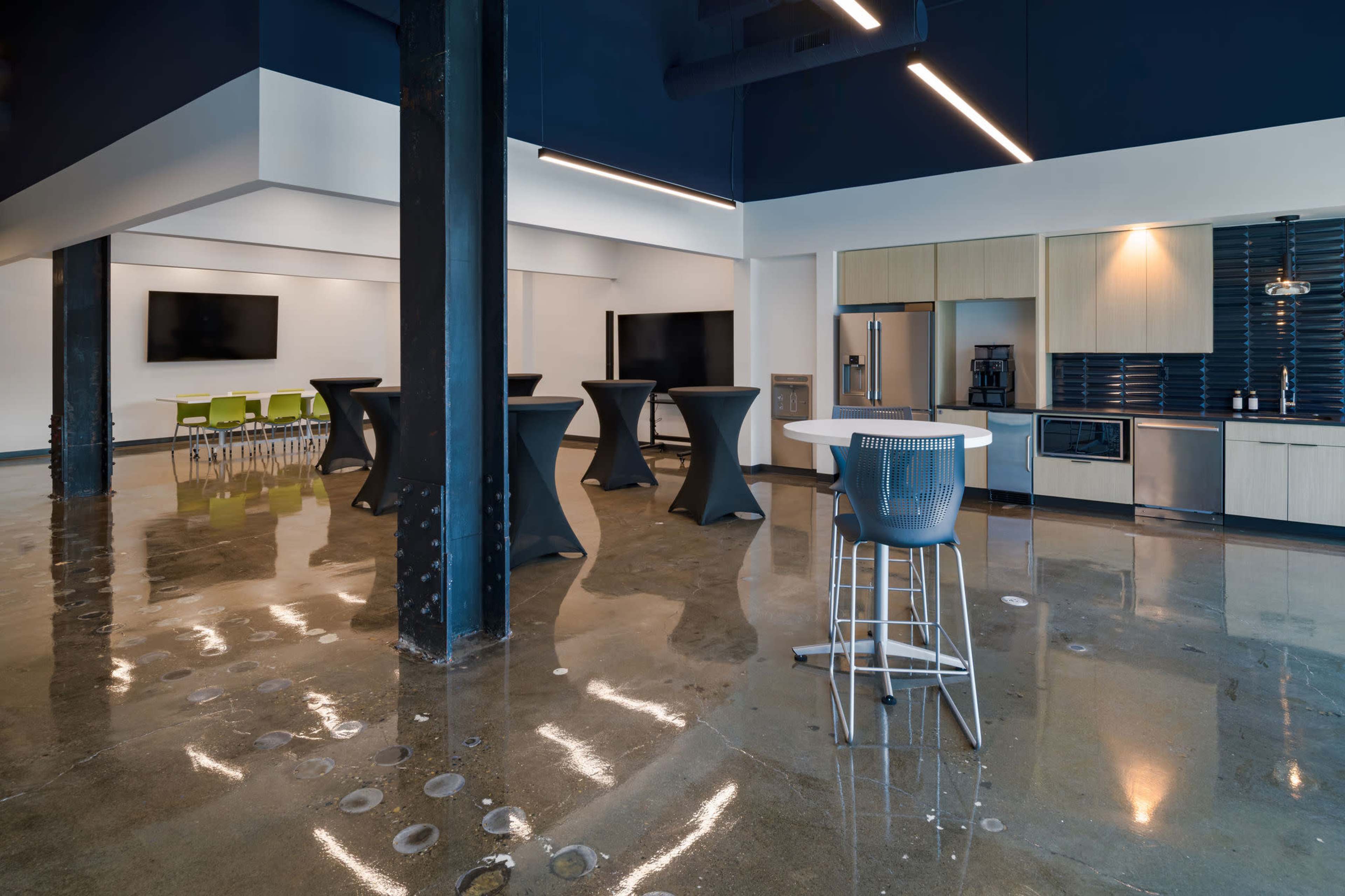 The image shows a modern interior space with polished concrete floors, featuring a kitchenette, tables, and a lounge area with seating.
