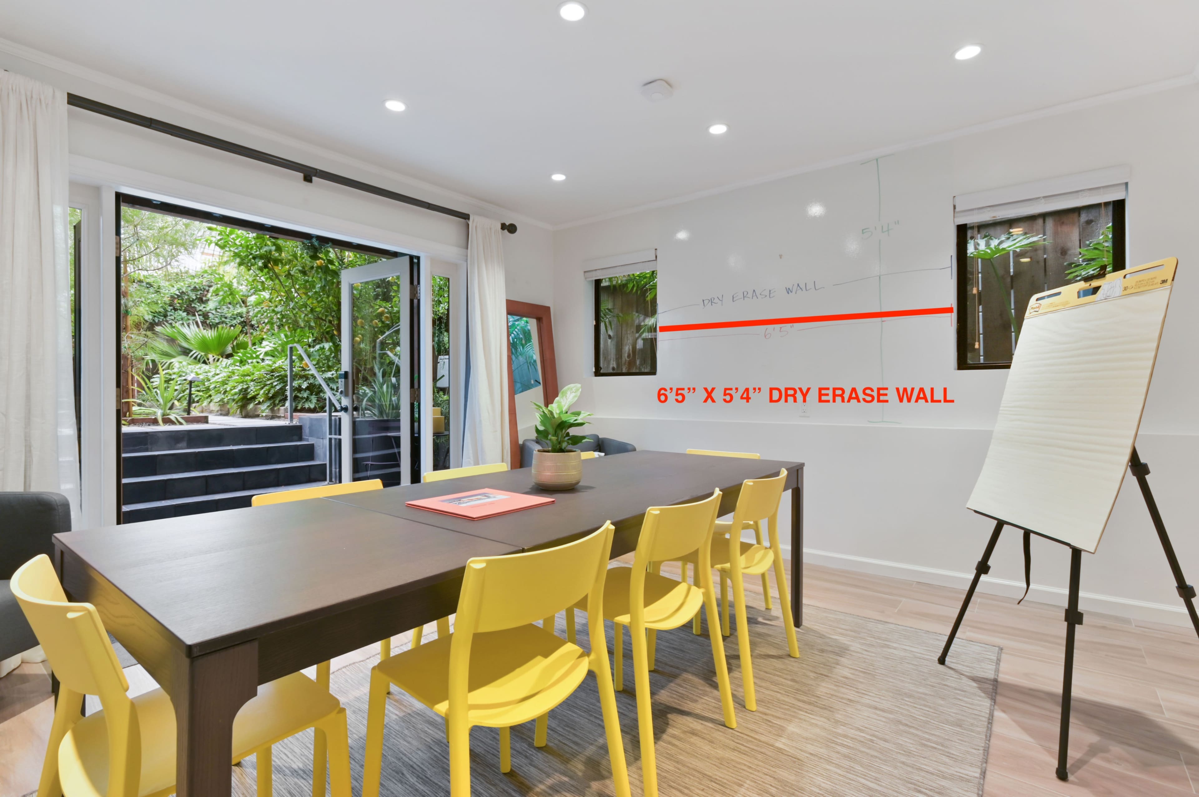 A conference room features a wooden table surrounded by yellow chairs, with a large dry erase wall and a view of an outdoor garden.