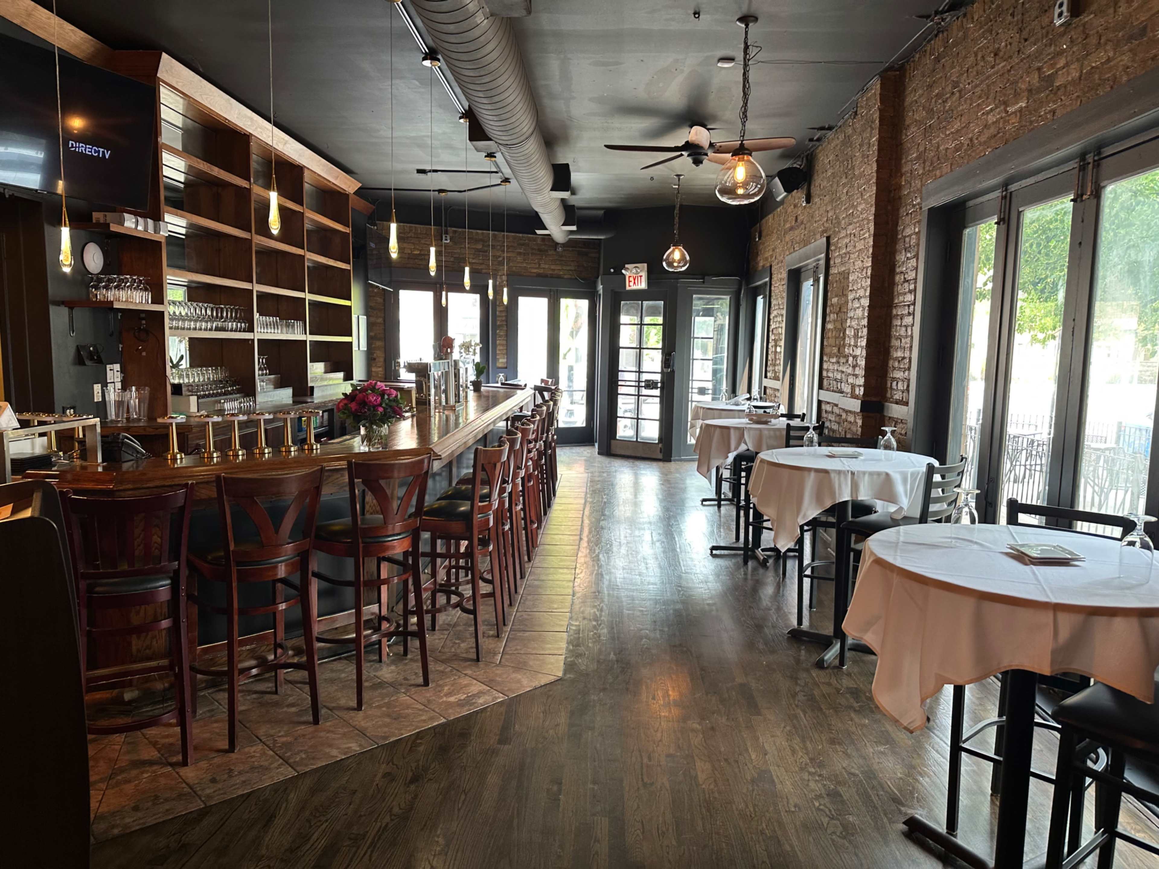 The image shows the interior of a restaurant featuring a bar area with wooden stools, a large window letting in light, and tables set for dining.