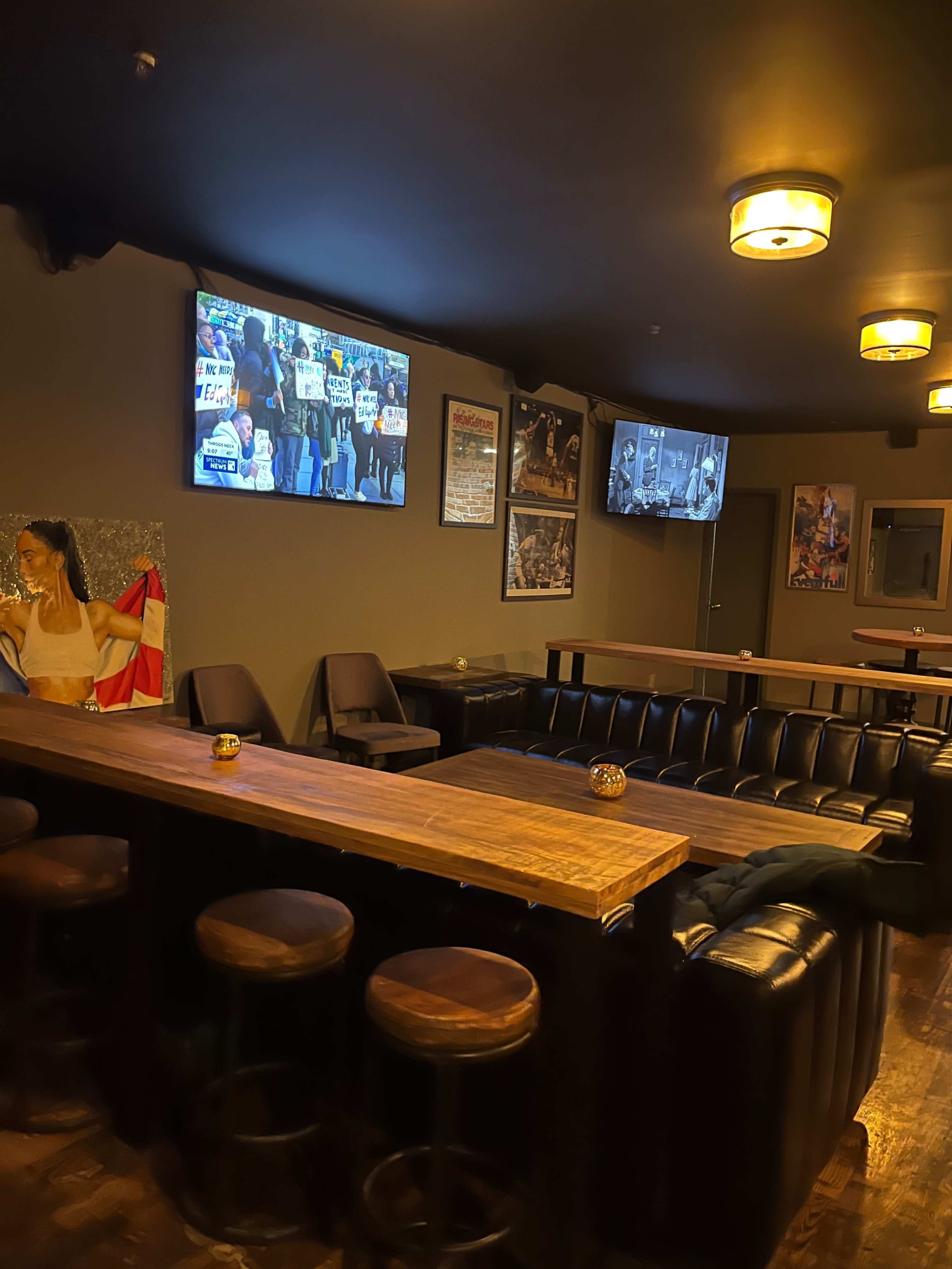 A dimly lit bar area with seating, wooden tables, and two screens displaying a protest scene.