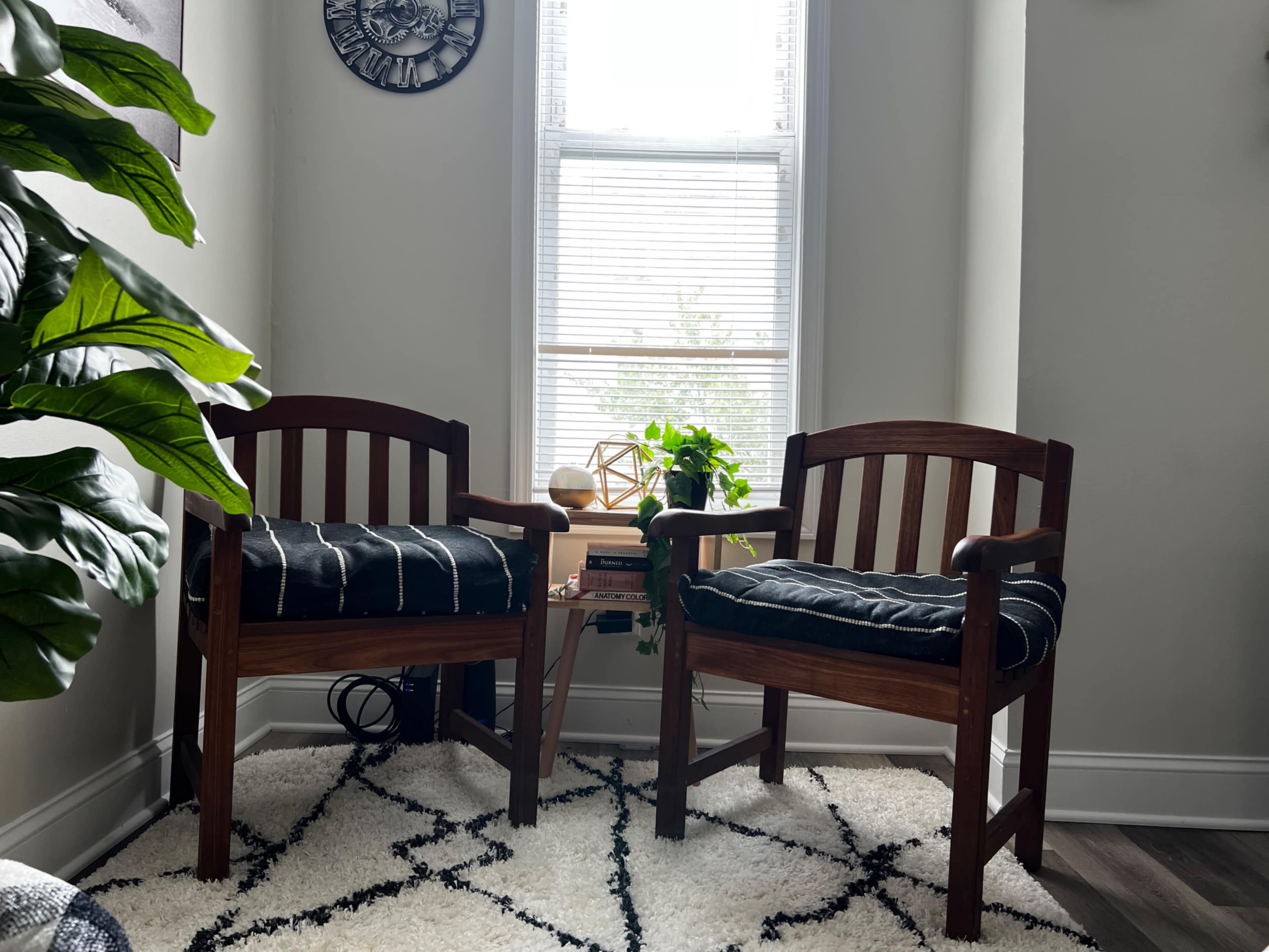 Two wooden chairs with cushions are positioned by a window surrounded by decorative plants and a small side table.