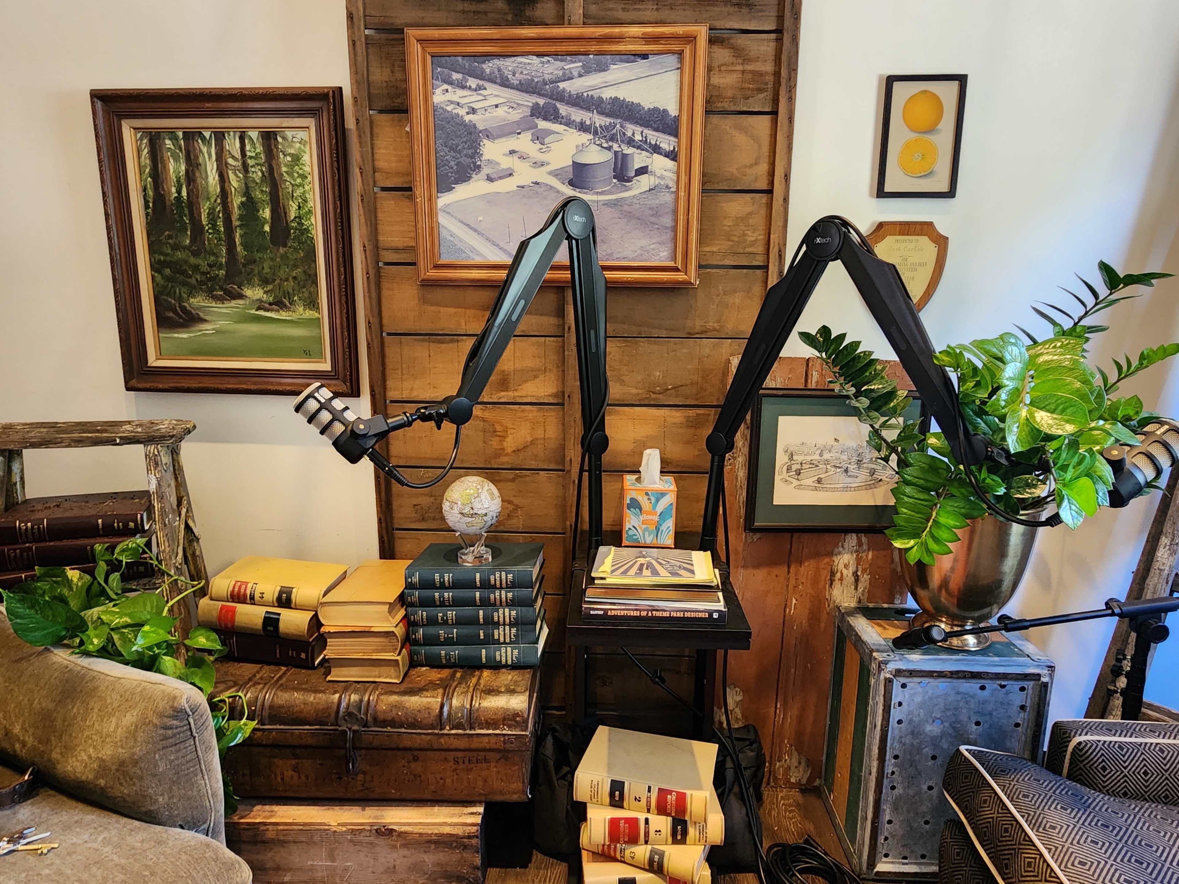 The image shows a corner of a room featuring a mix of framed artwork, an antique trunk, stacks of books, and two large articulated microphone arms positioned near a small table with a decorative object.
