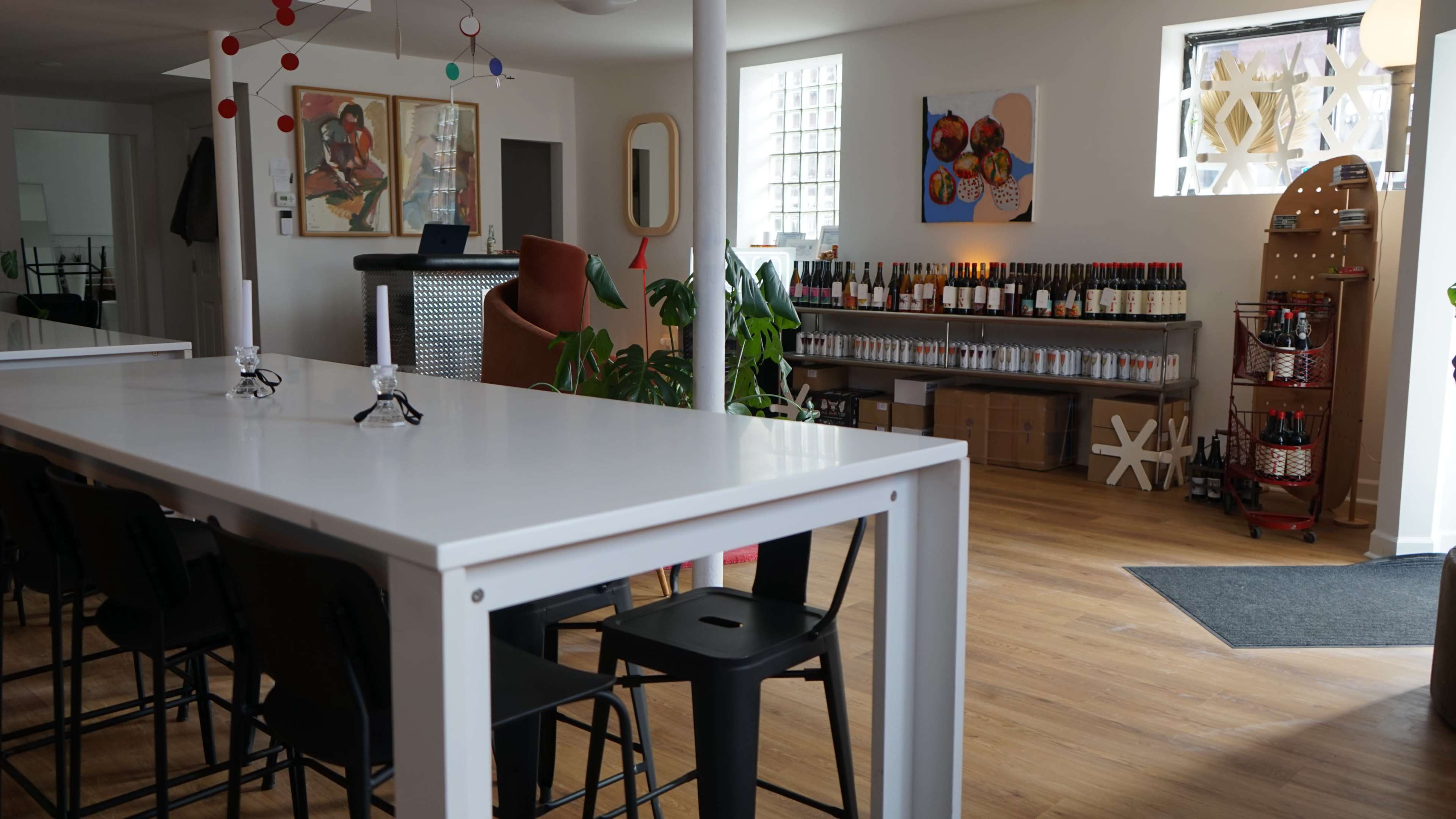 The image shows a modern interior space featuring a long white table with black chairs, a bar area in the background, and shelves filled with bottles along the walls.