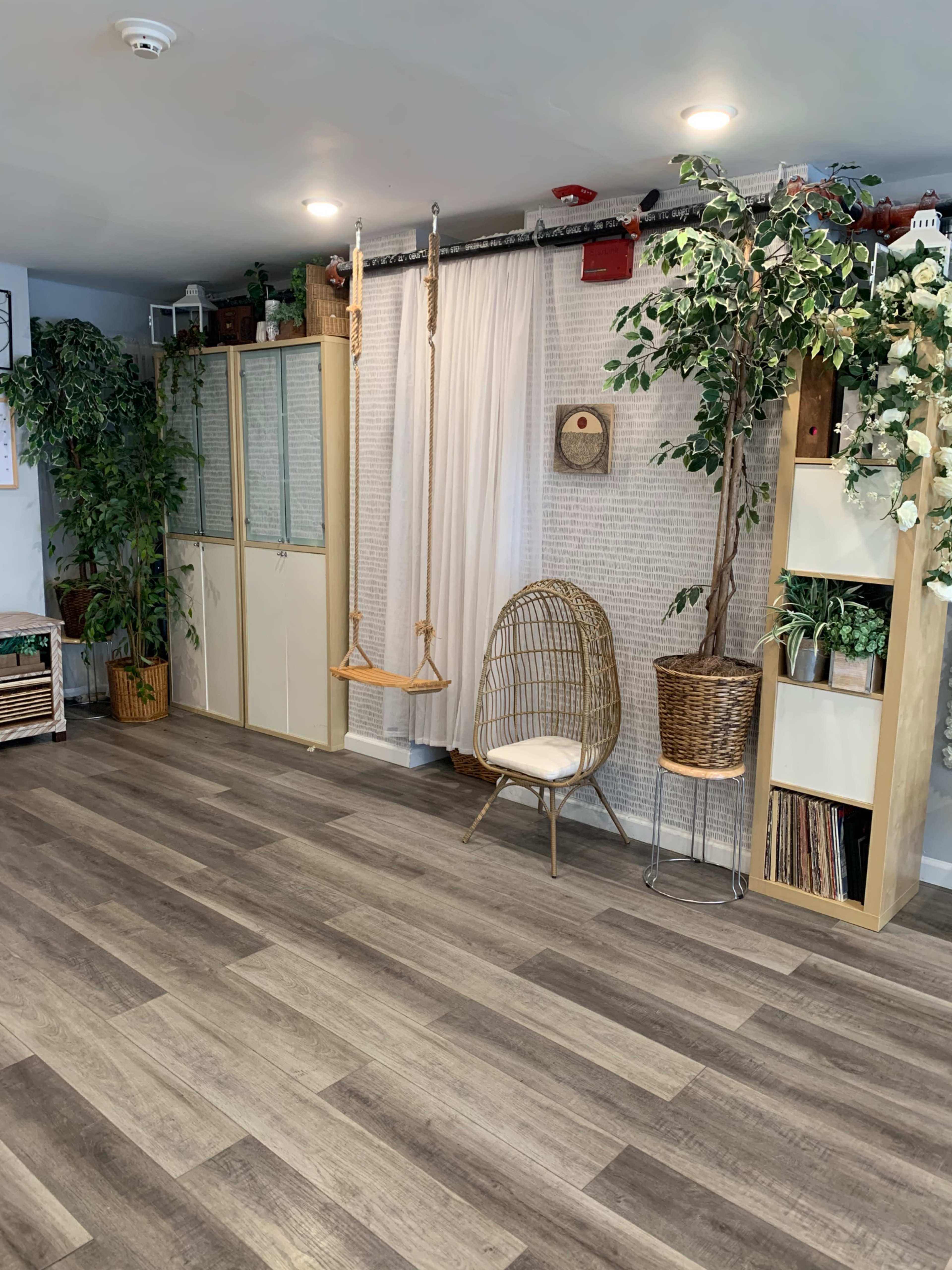 The room features a swing hanging from the ceiling, plants in various containers, a rattan chair, and shelving units with decor and storage.