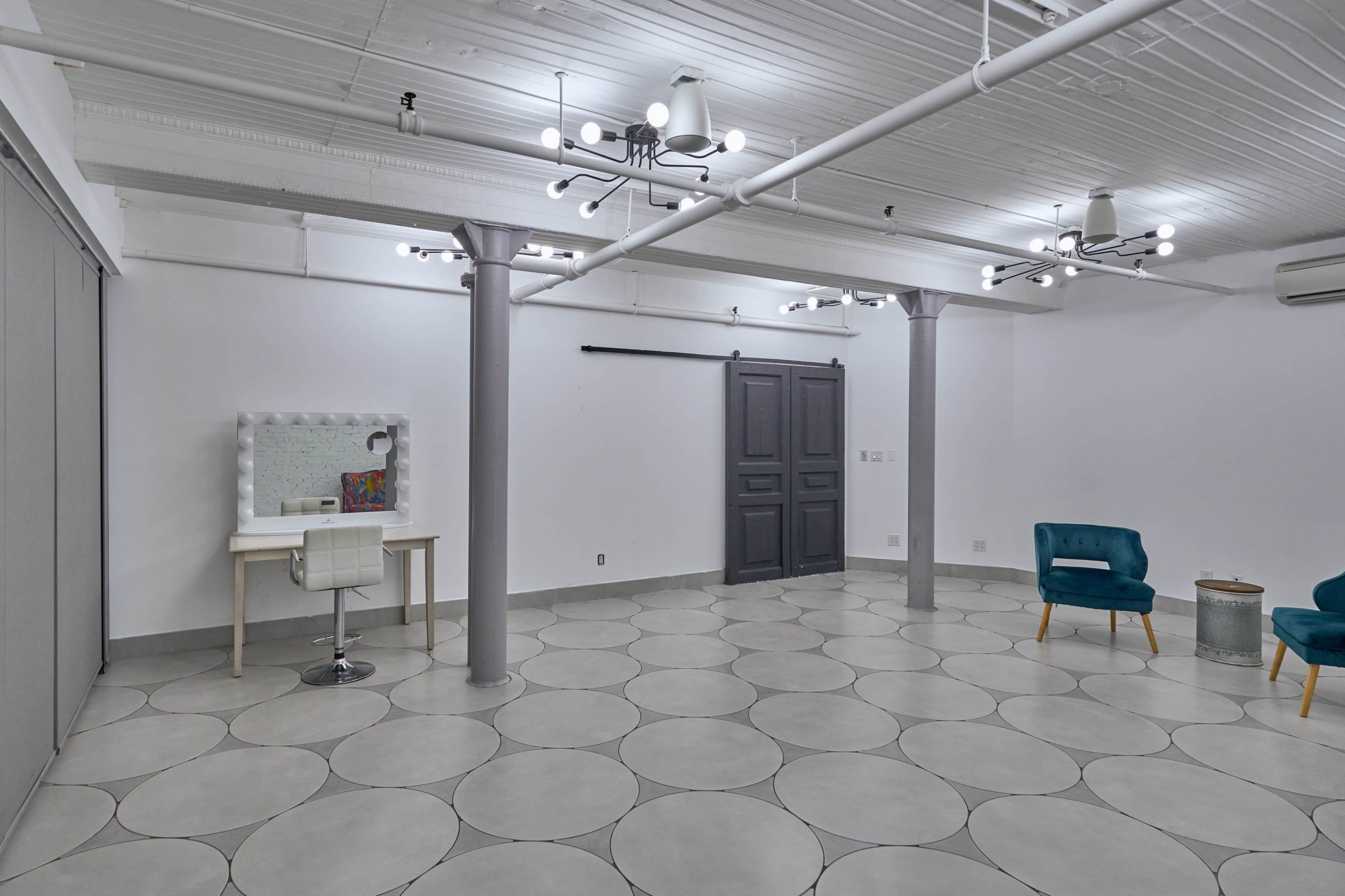 A spacious, well-lit room with a simple gray floor featuring circular patterns, a vanity with a mirror, and two teal chairs.