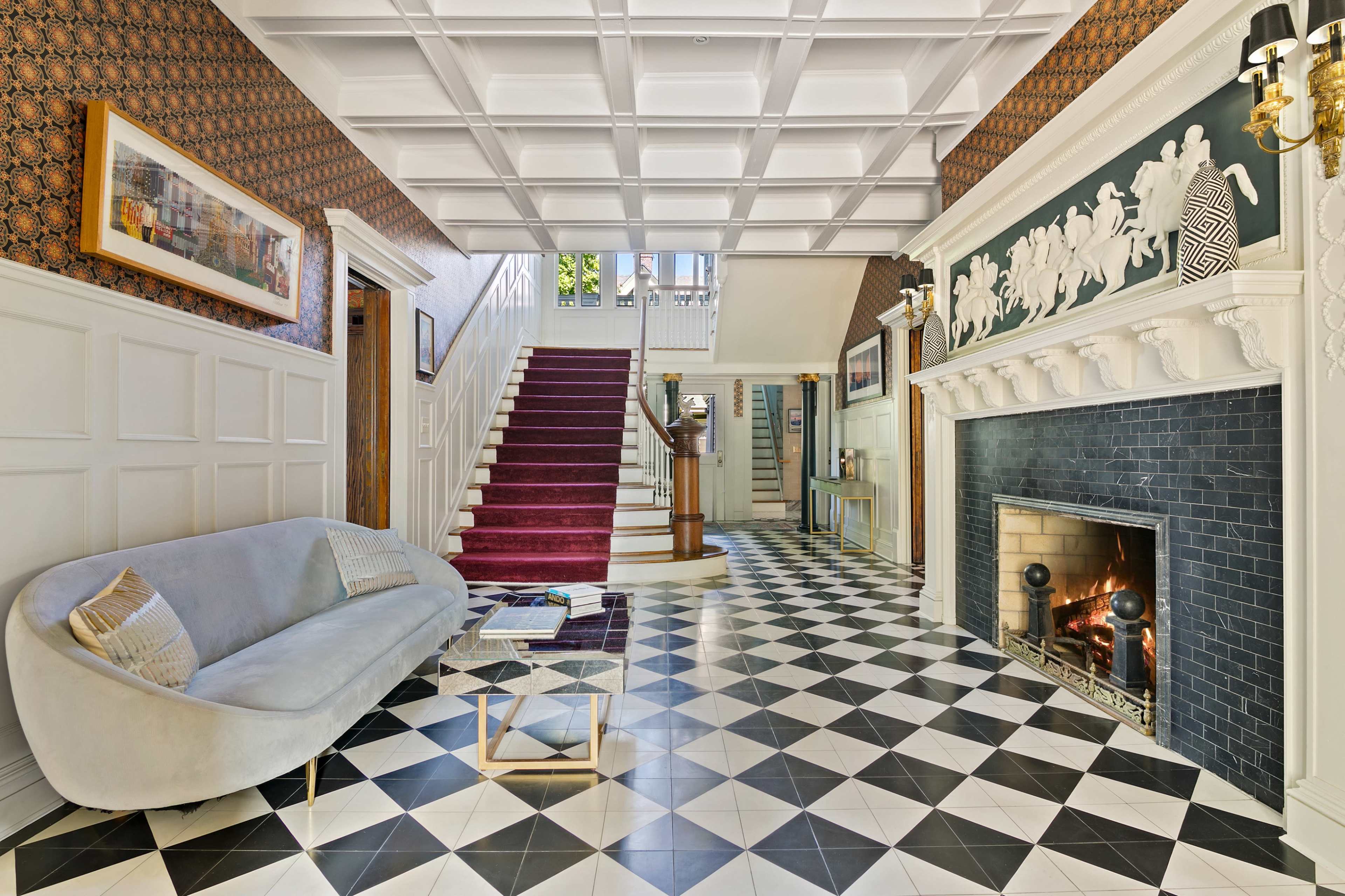 The image shows an elegantly designed foyer featuring a patterned floor, a staircase, a fireplace, and a modern sofa.