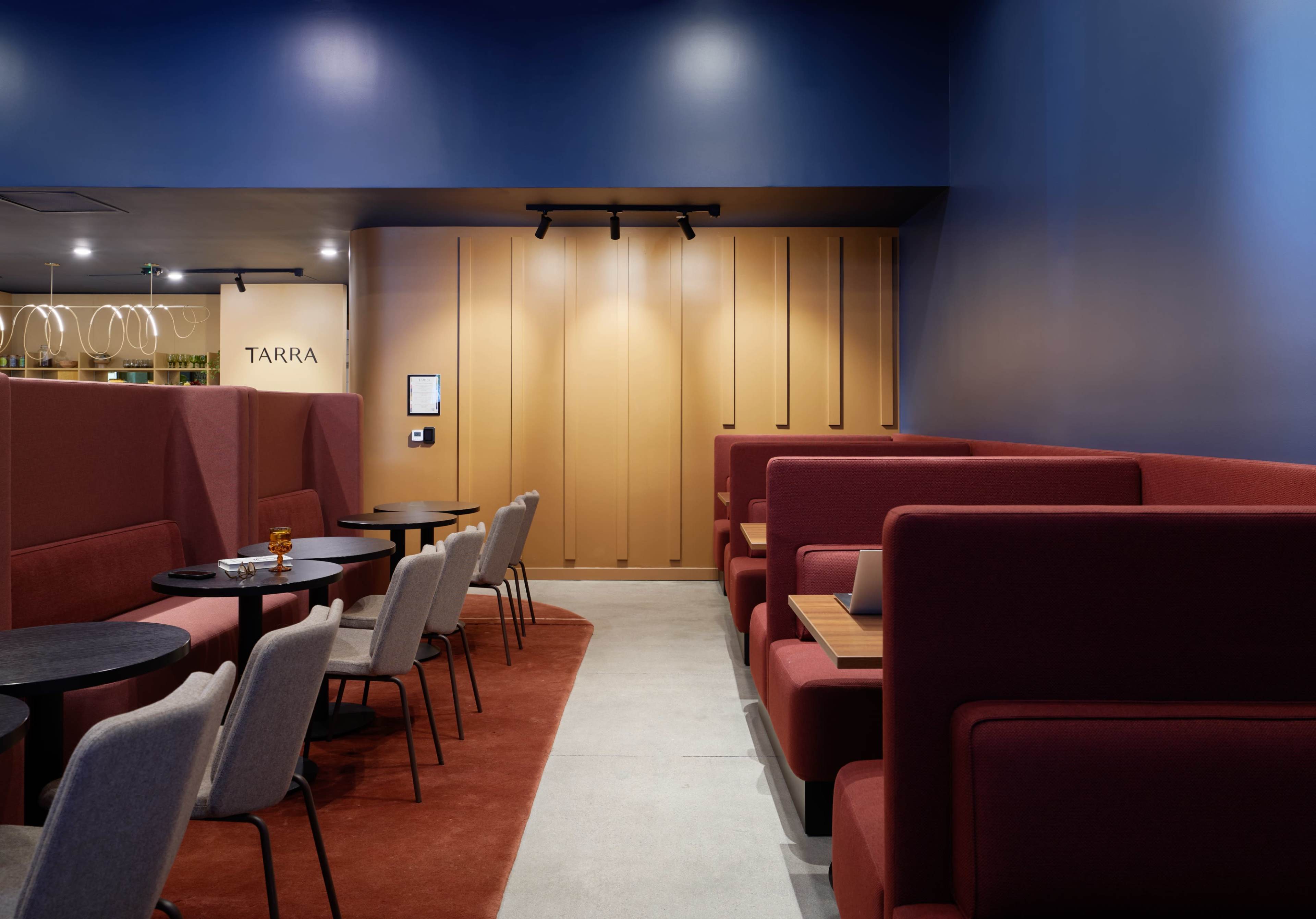 The image shows a modern café interior featuring red seating booths, gray chairs, and a blue-painted wall, creating a cozy atmosphere.
