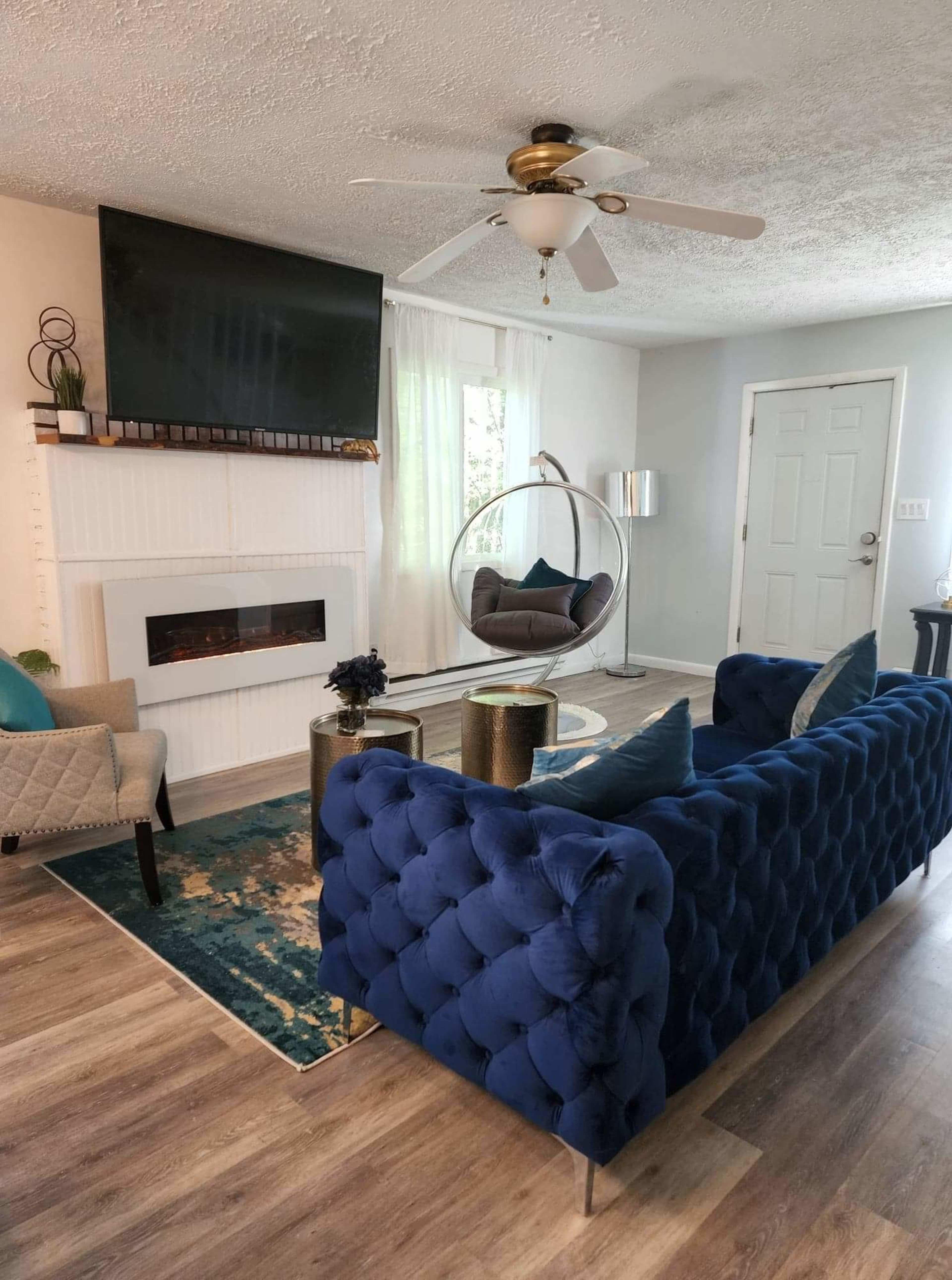 The living room features a blue tufted couch facing a modern TV above a white fireplace, with a circular chair and decorative side tables nearby.