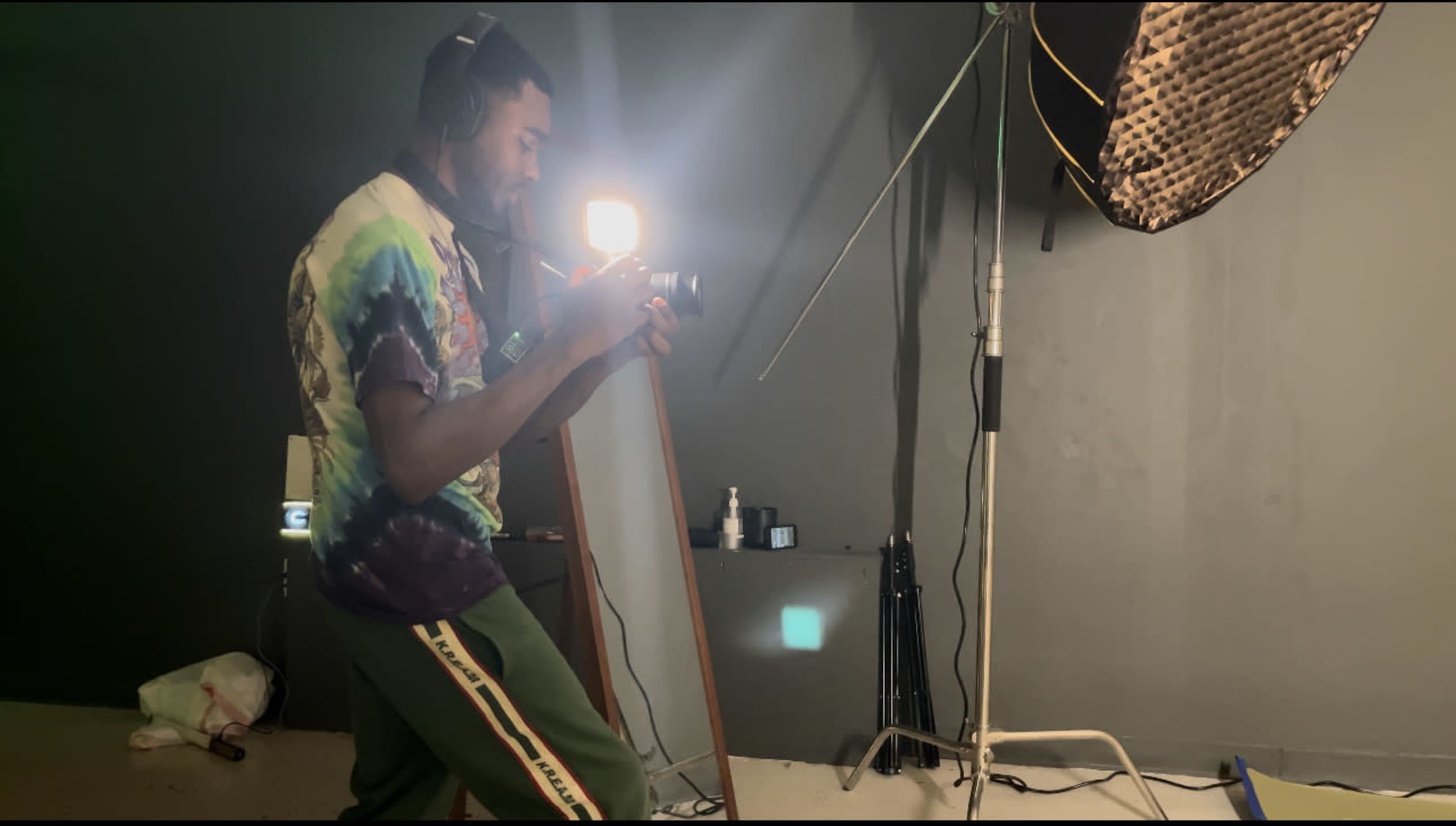 A man wearing a tie-dye shirt uses a camera while standing in a photography studio with lighting equipment.