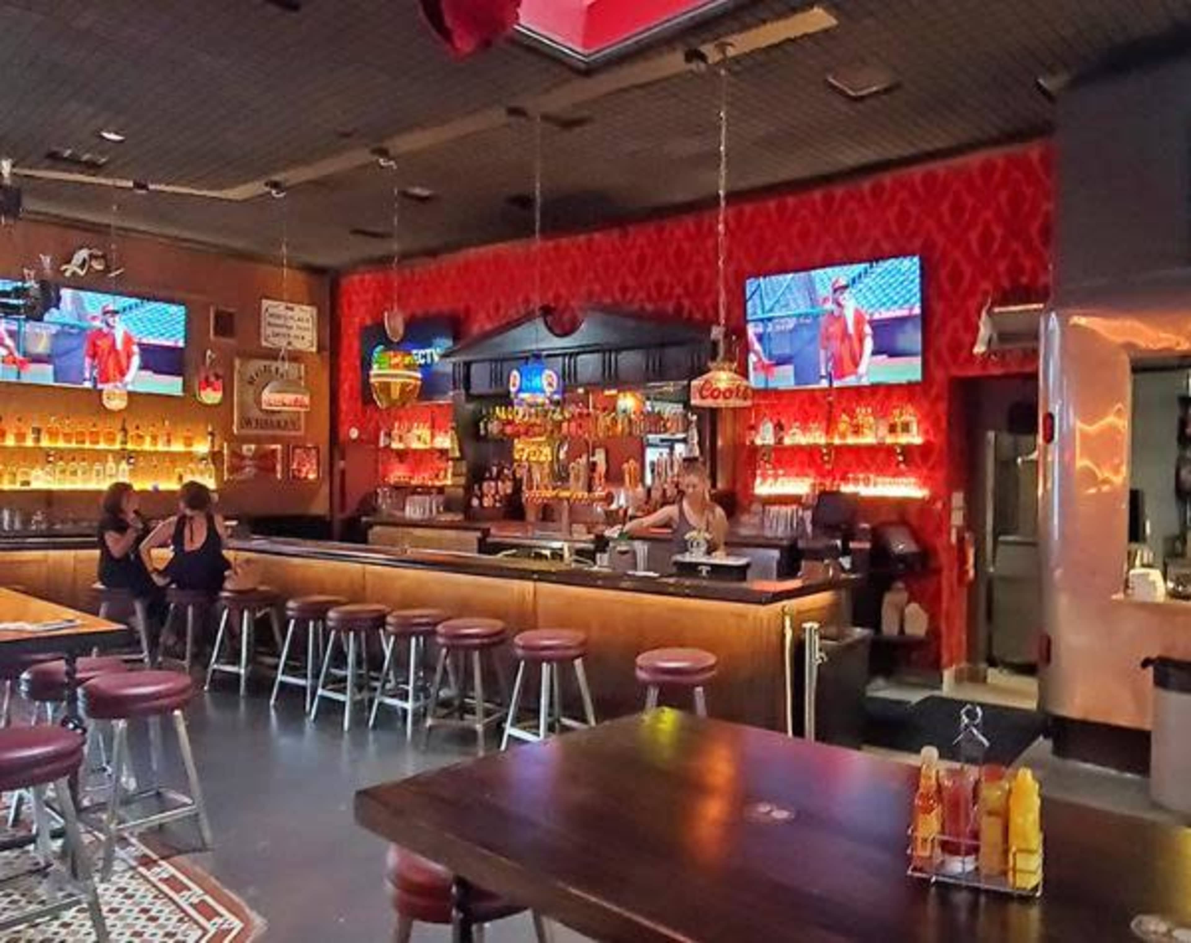 A bar area featuring a long countertop, high stools, two televisions, and a bright red decorative wall with various beverage displays.