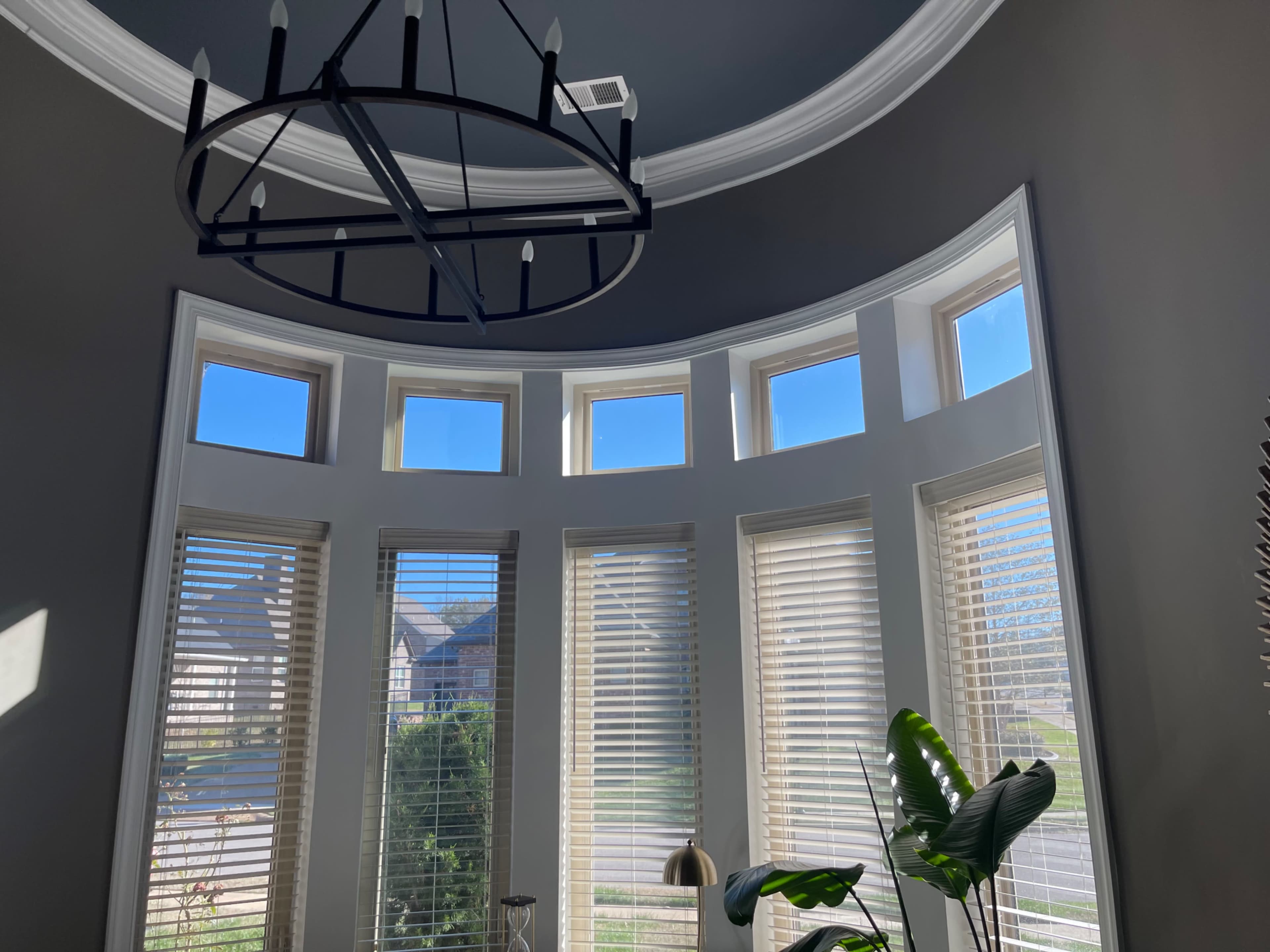 A room with a large circular chandelier and multiple bay windows featuring white blinds, allowing natural light to brighten the space.