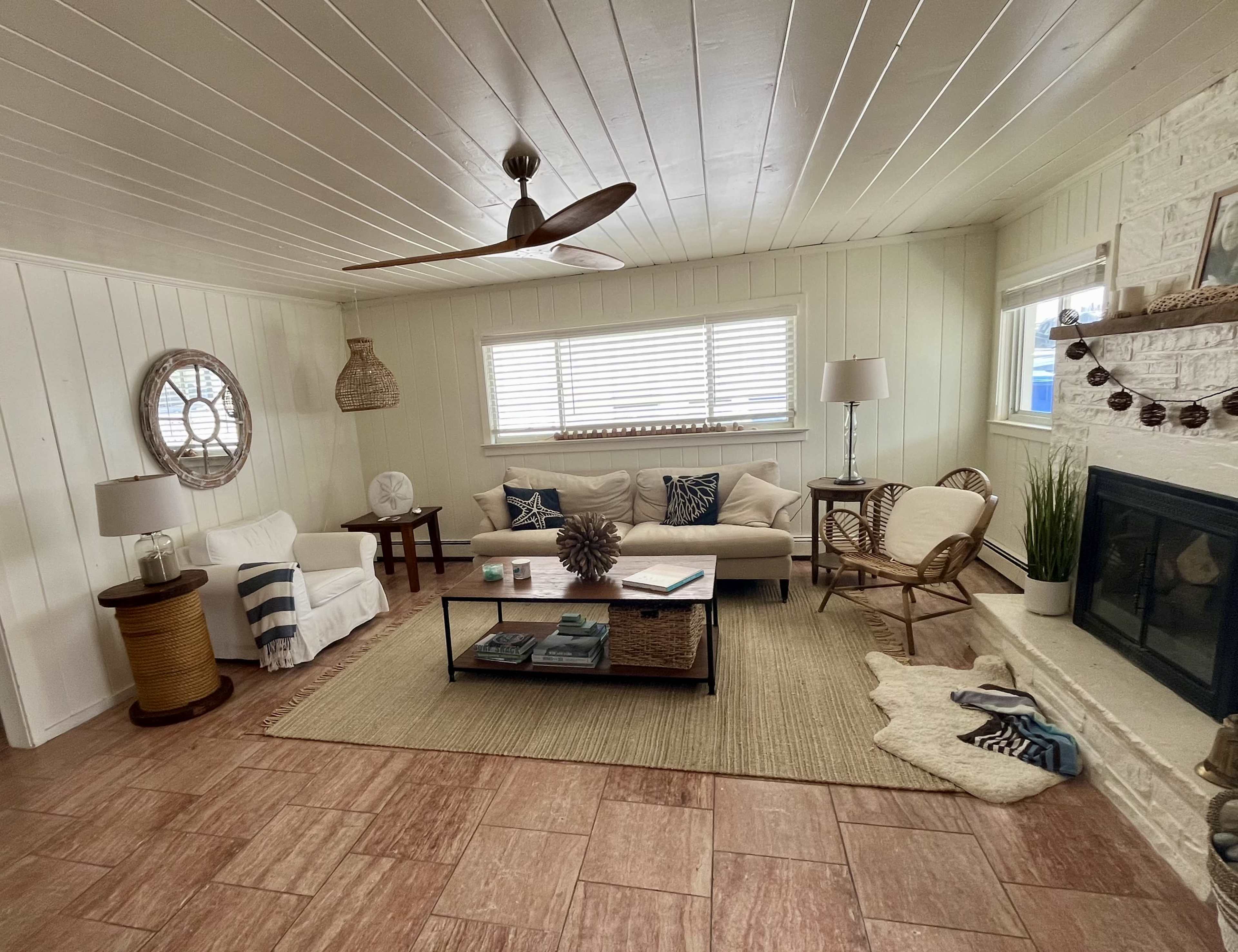 The living room features a light-colored sofa, a coffee table with books, a woven rug, and a fireplace surrounded by natural light from several windows.
