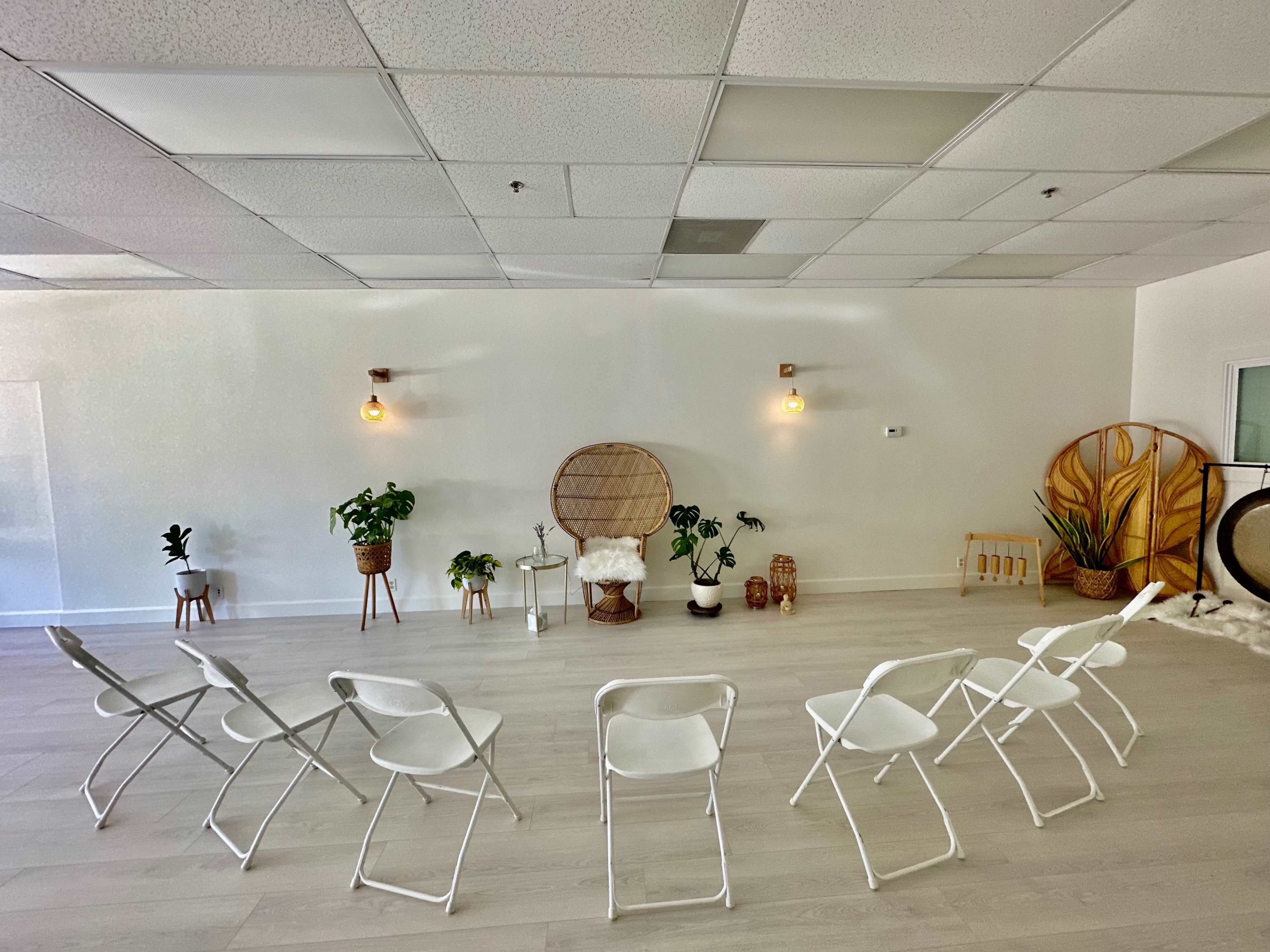 A minimalist room features white folding chairs arranged in a semicircle facing a decorative backdrop with plants and soft lighting.