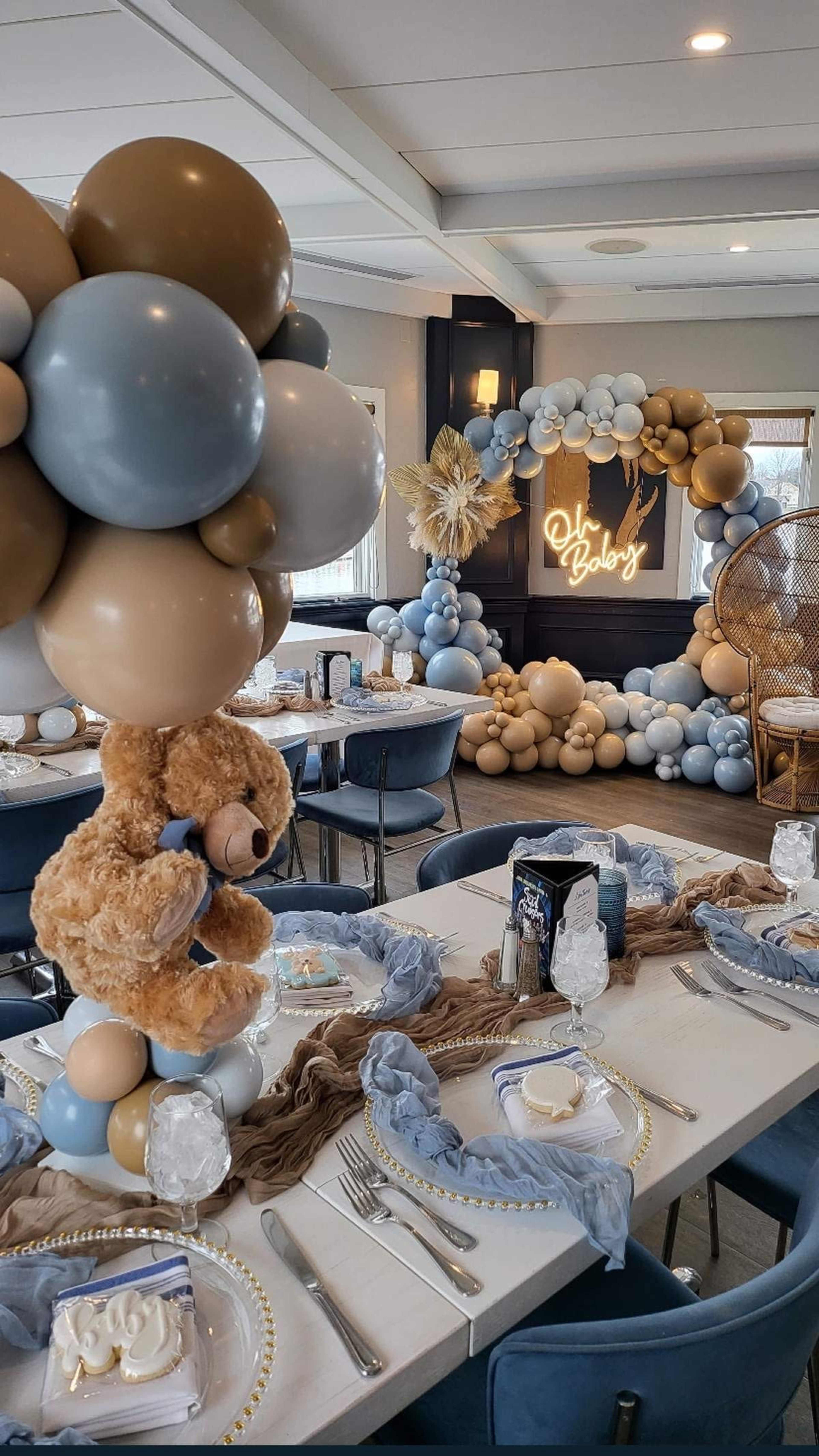 The scene depicts a party setup featuring tables adorned with light blue and beige decorations, elegant tableware, and balloon arrangements, including a teddy bear centerpiece and a backdrop with illuminated text.