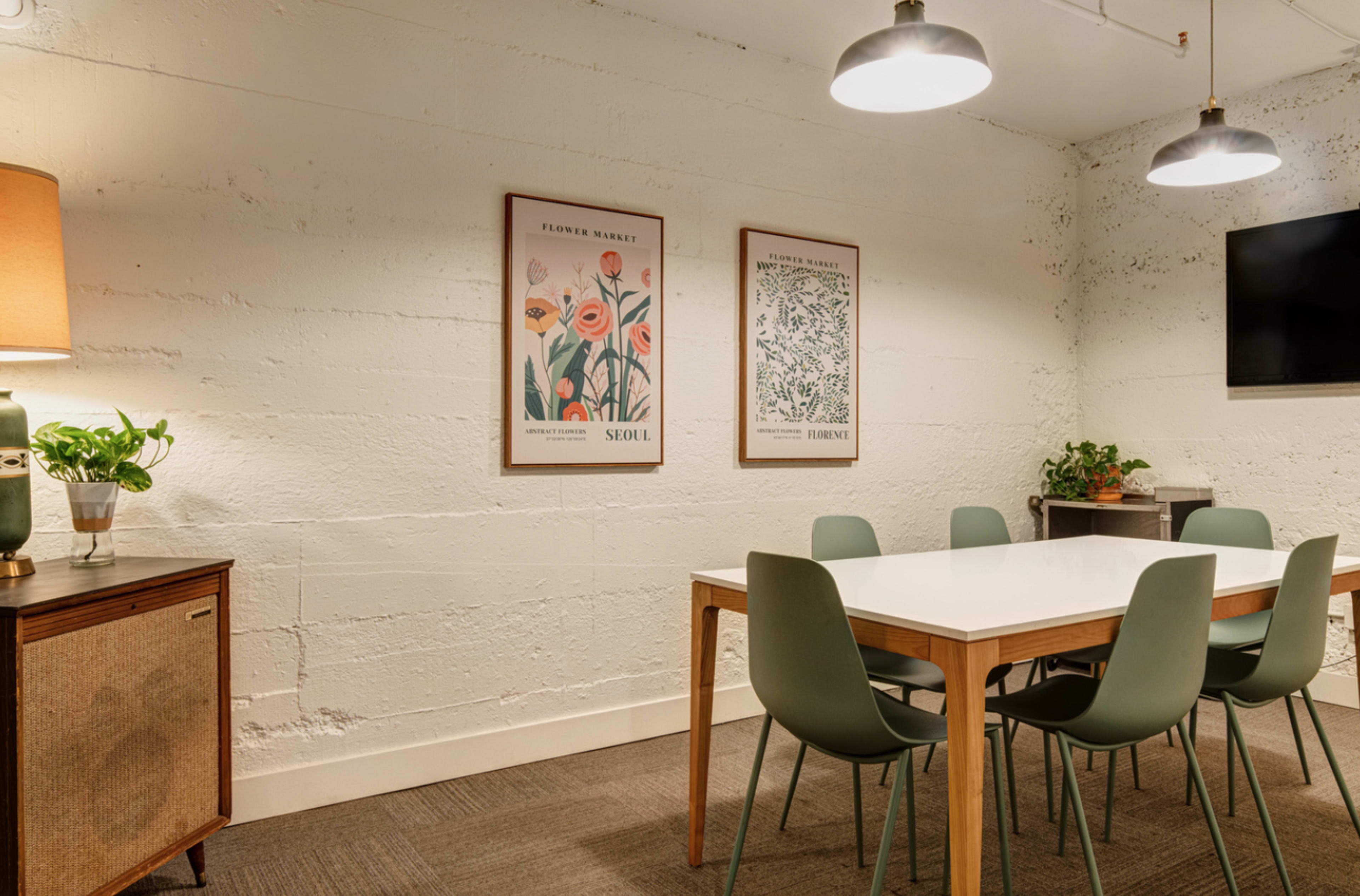 A modern meeting room features a white table surrounded by green chairs, with framed artwork on the walls and a television in the corner.