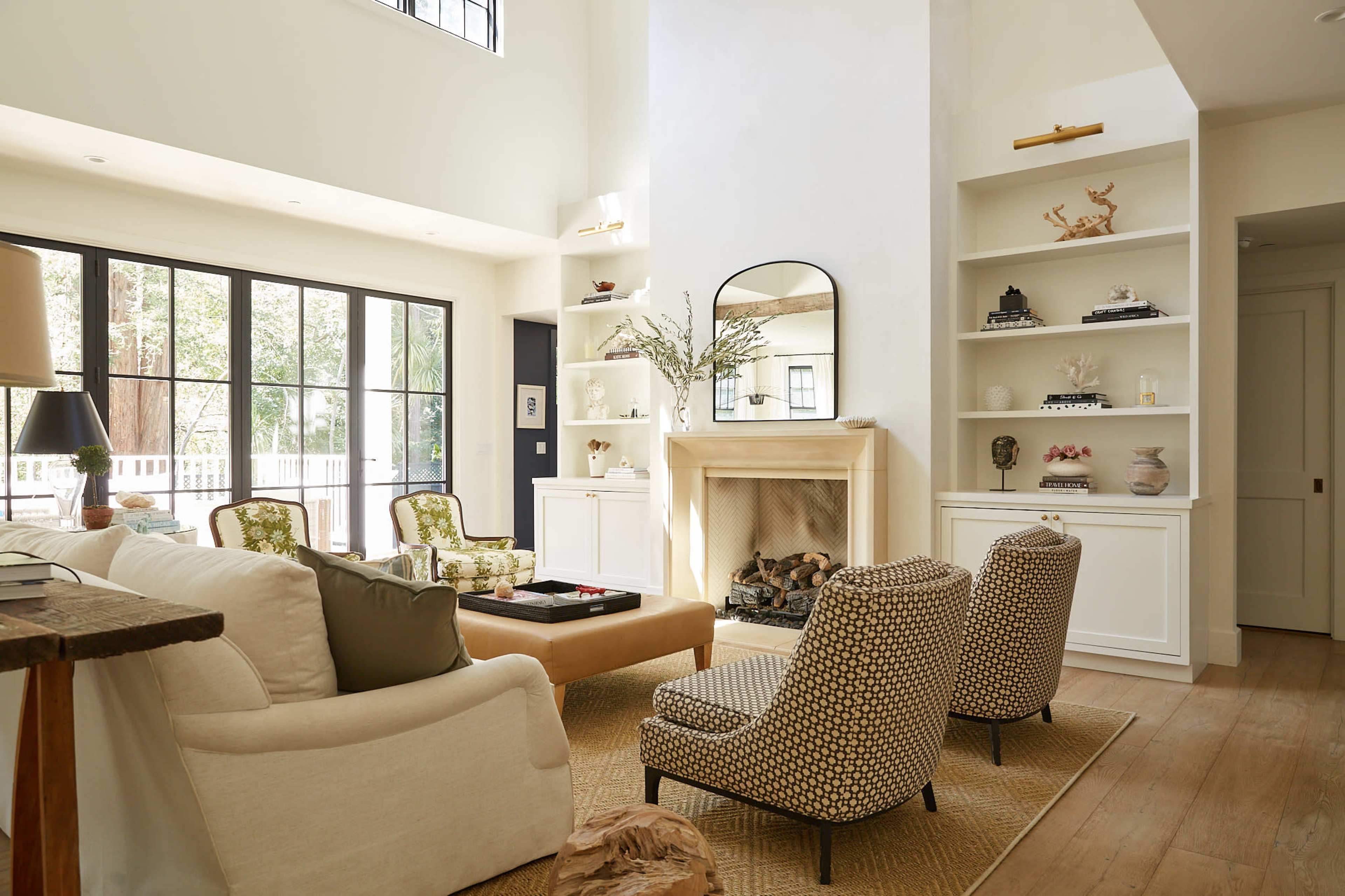 A bright living room with a large window, a fireplace, built-in shelves, and two patterned armchairs next to a neutral sofa.