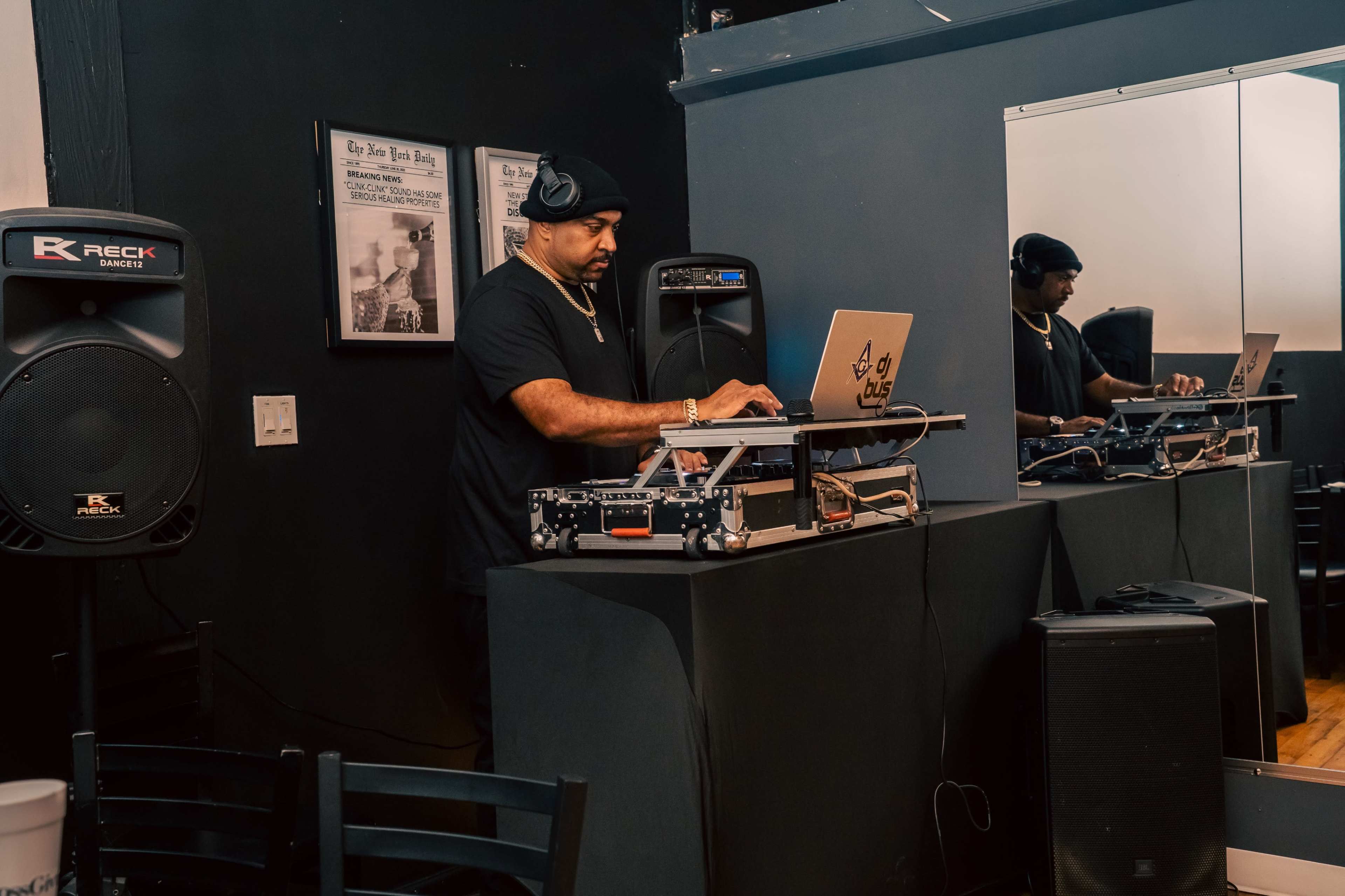 A DJ is mixing music behind a laptop and turntables in a small, dimly lit venue with black walls and a mirror.