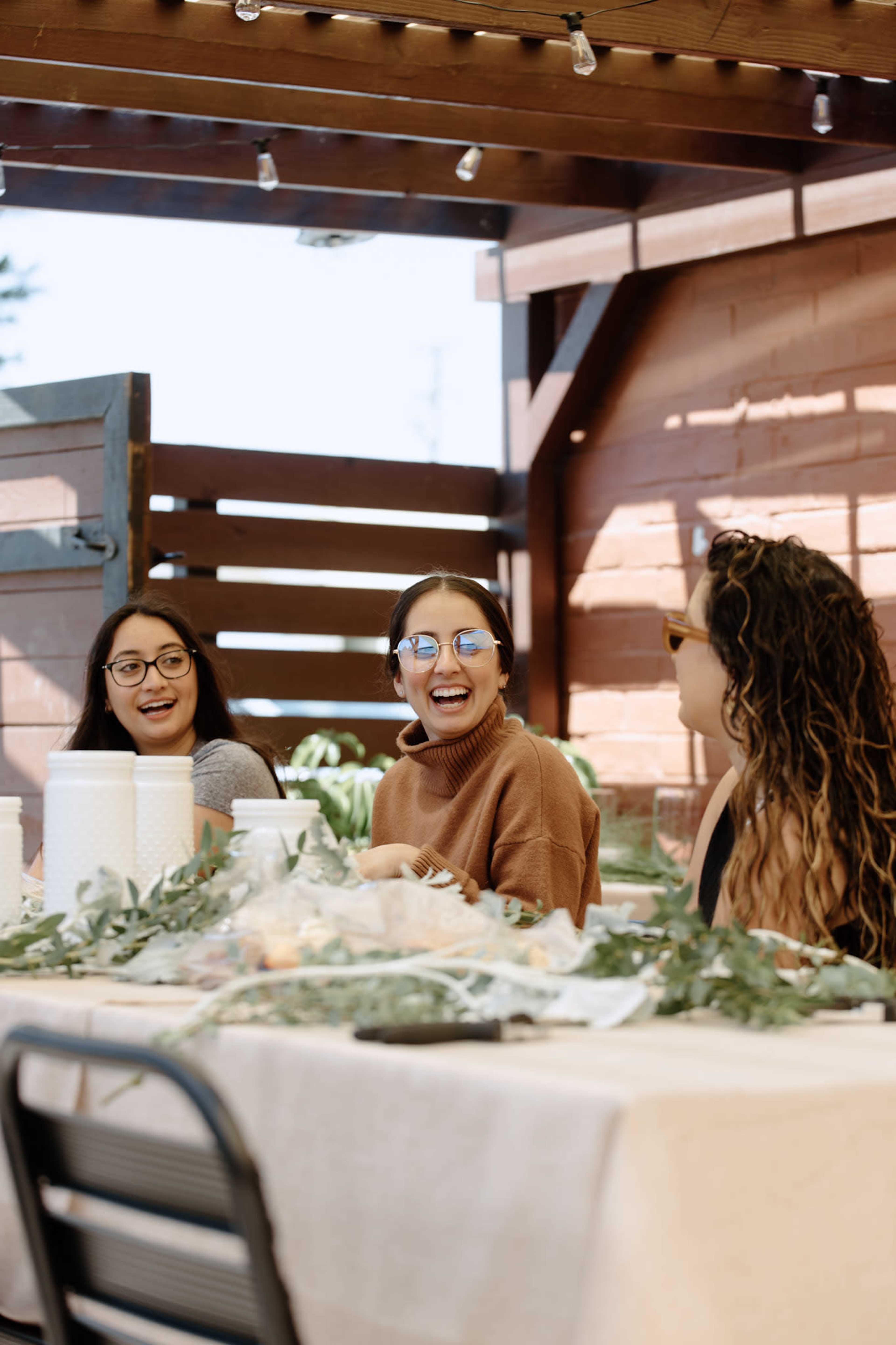 Three women are seated at a table adorned with greenery and crafting supplies, engaging in conversation under a wooden structure.