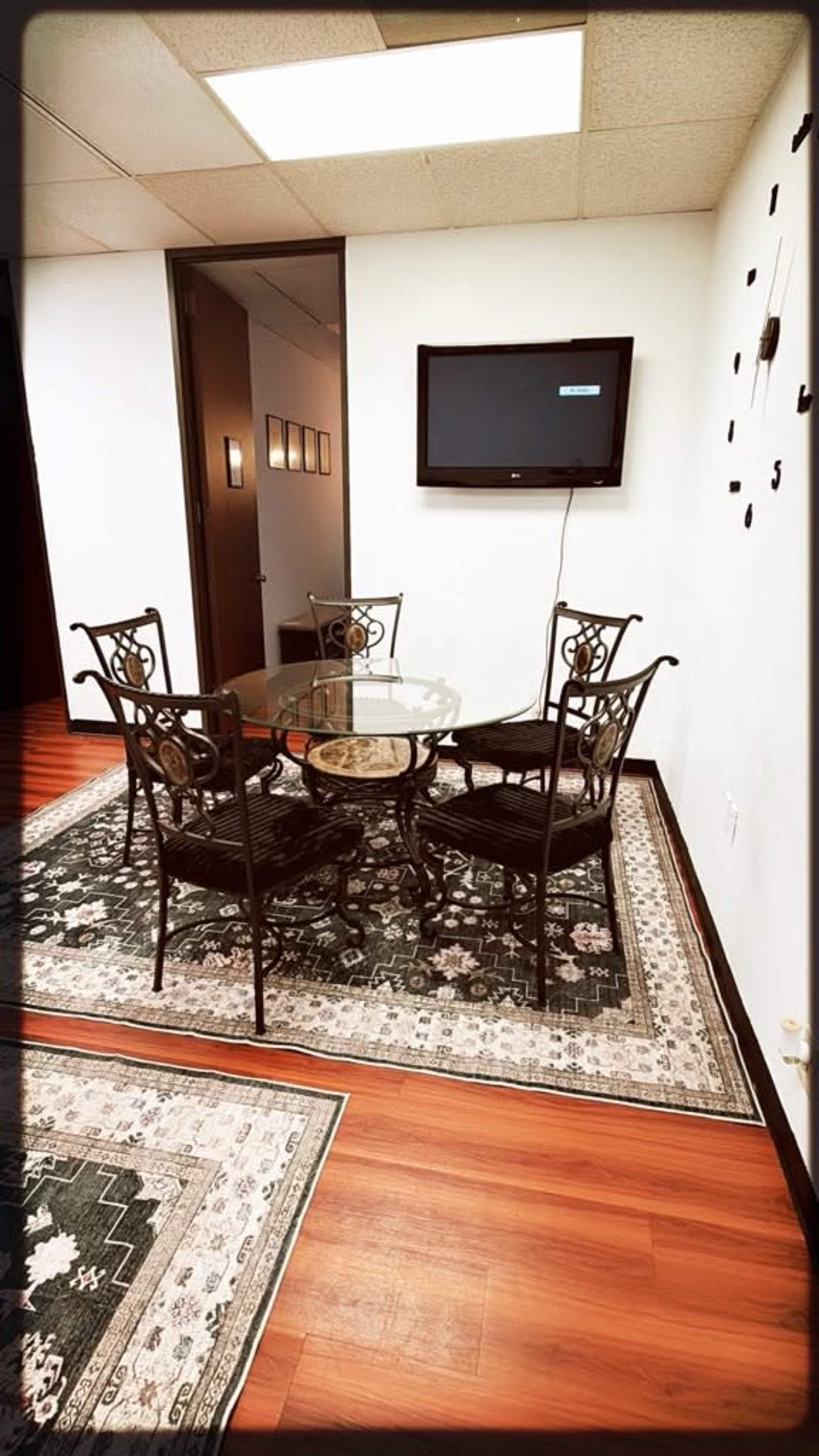 The image shows a small meeting room with a glass dining table and four black iron chairs on a patterned rug, accompanied by a wall-mounted television.