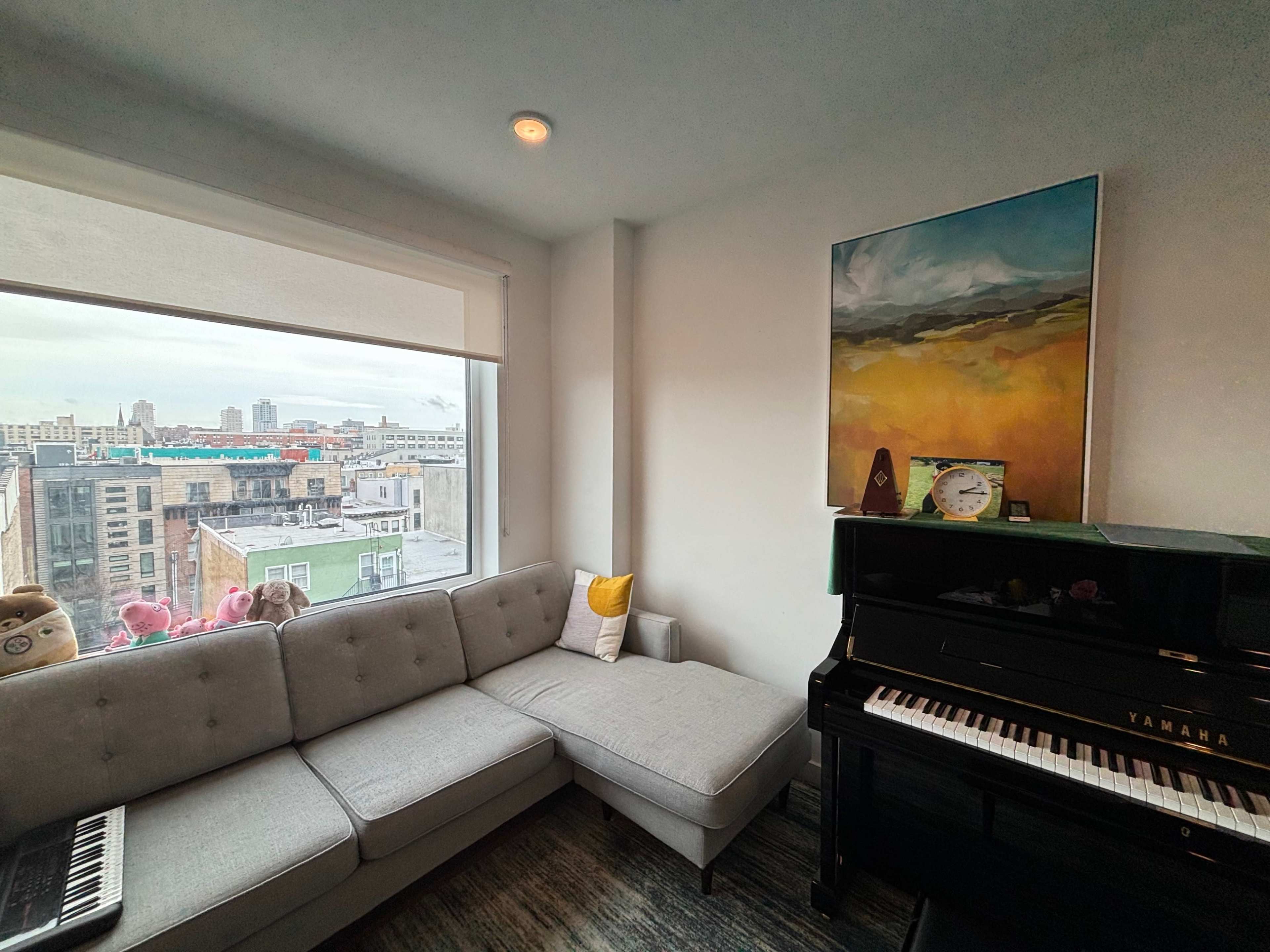 A corner of a room features a gray sectional couch, a piano, and a window showcasing a cityscape with buildings.