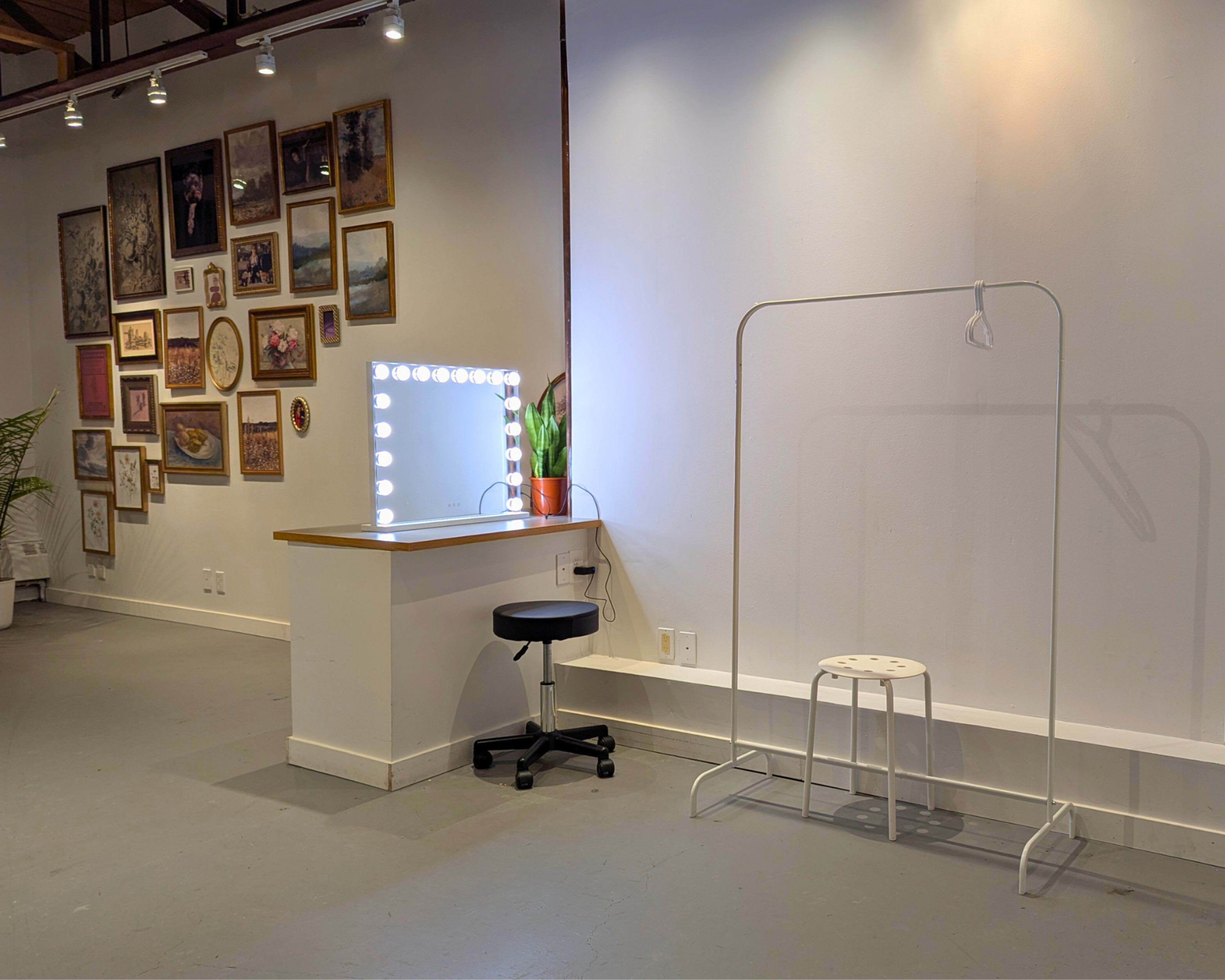 The image shows an interior space featuring a wall adorned with various framed pictures, a mirror with lights, a small desk with a stool, and a clothing rack.