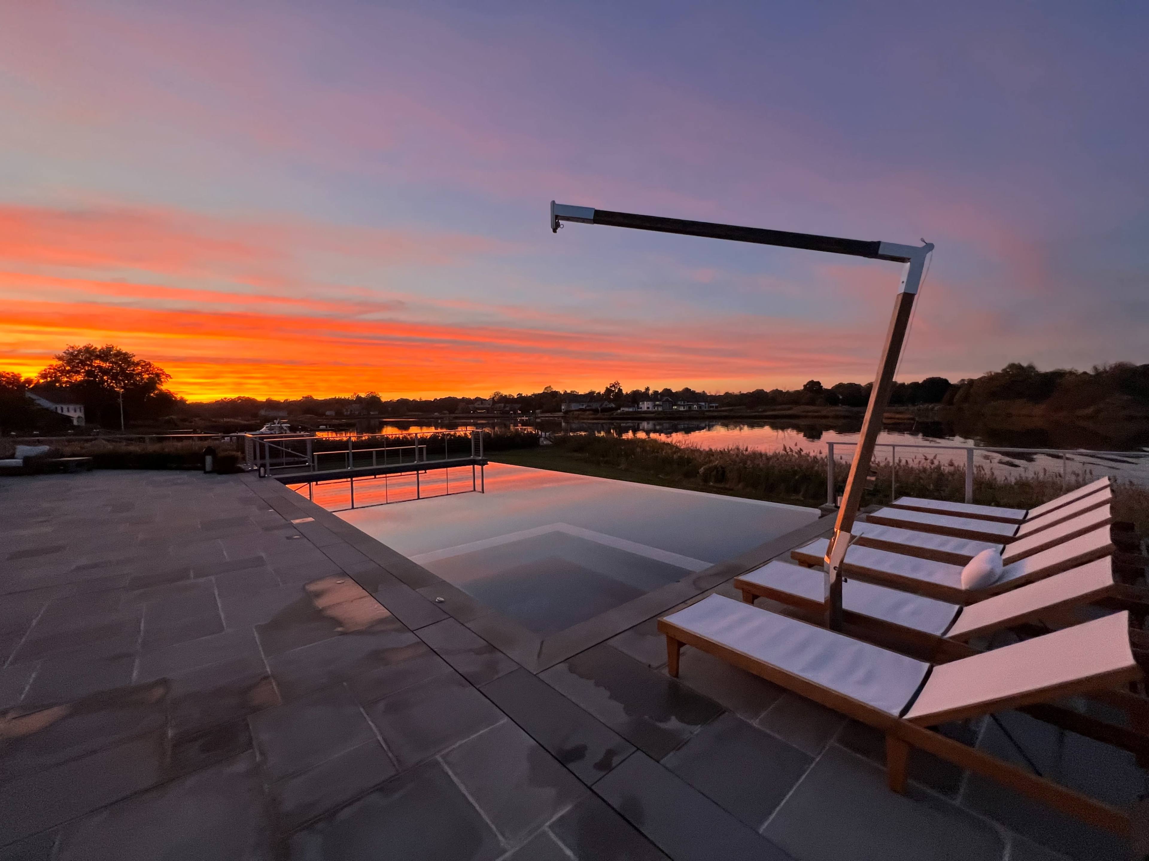 A poolside setting with lounge chairs beside a tranquil water surface reflecting a colorful sunset.