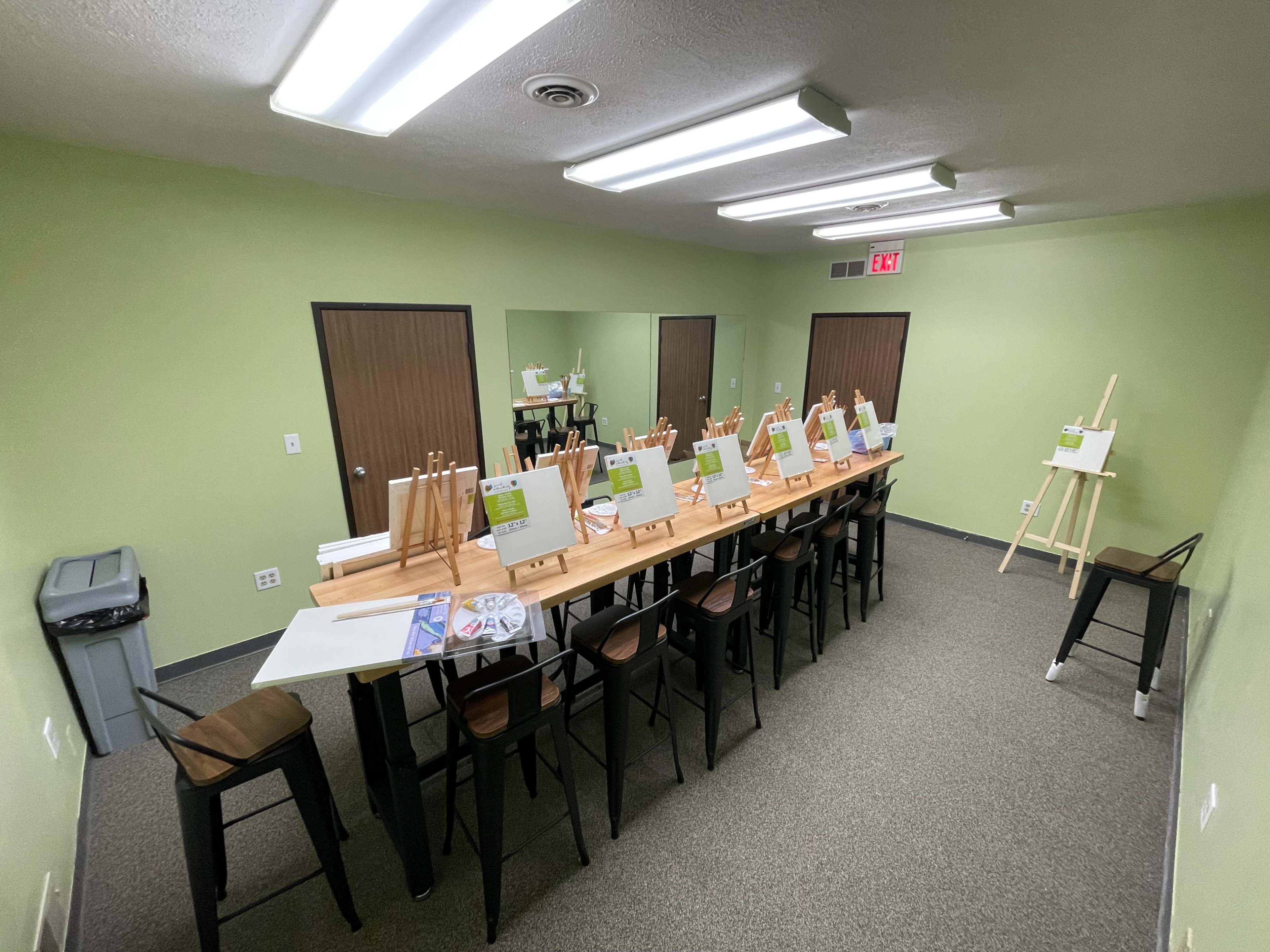 A spacious studio is set up with a long table lined with easels and blank canvases, surrounded by chairs and mirrors on the walls.