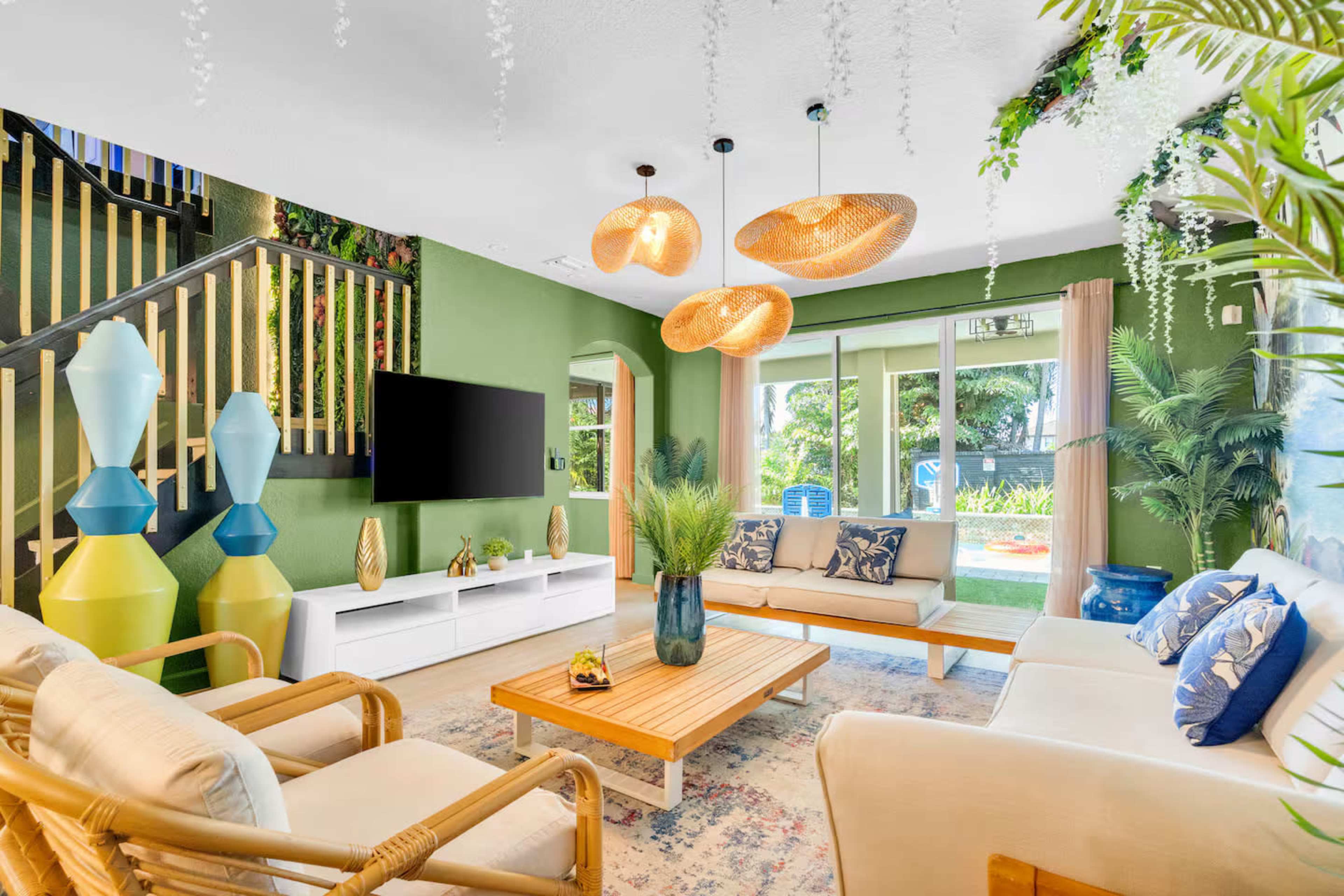 The living room features a modern design with rattan furniture, a wooden coffee table, green walls, and pendant lighting, along with a television mounted on the wall.