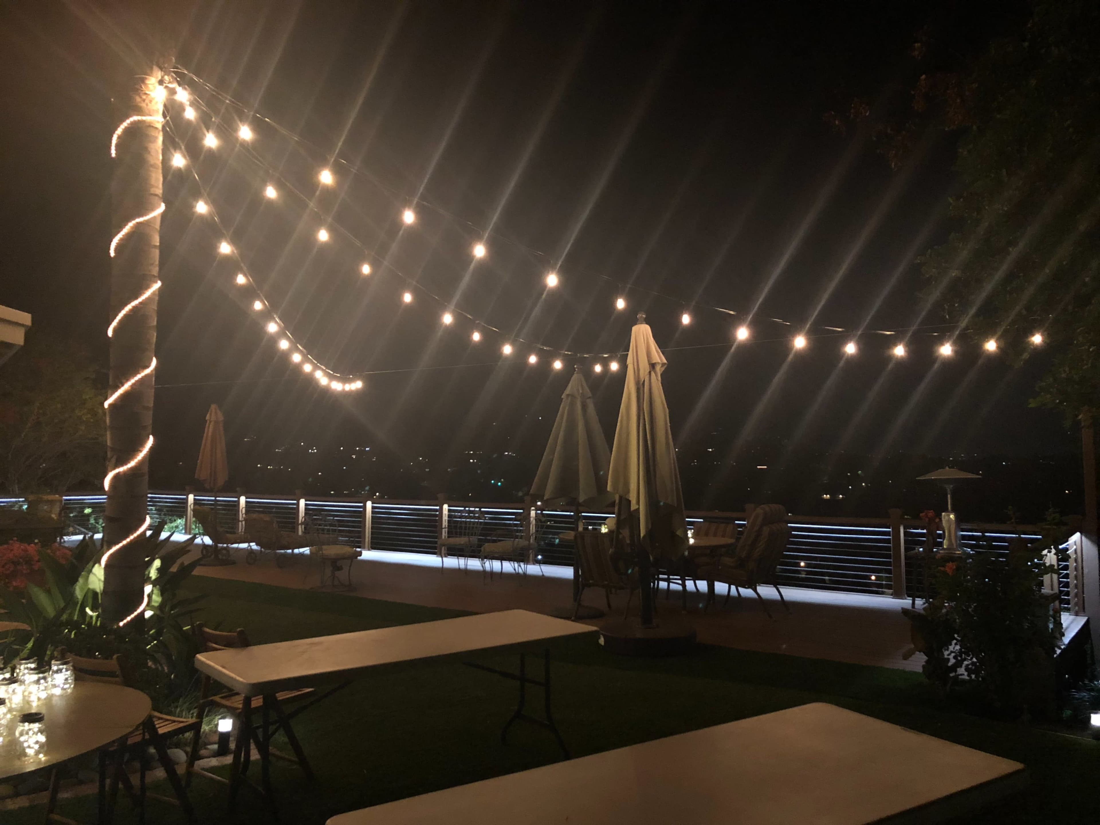 A nighttime outdoor space with string lights illuminating a patio area, featuring tables and umbrellas.