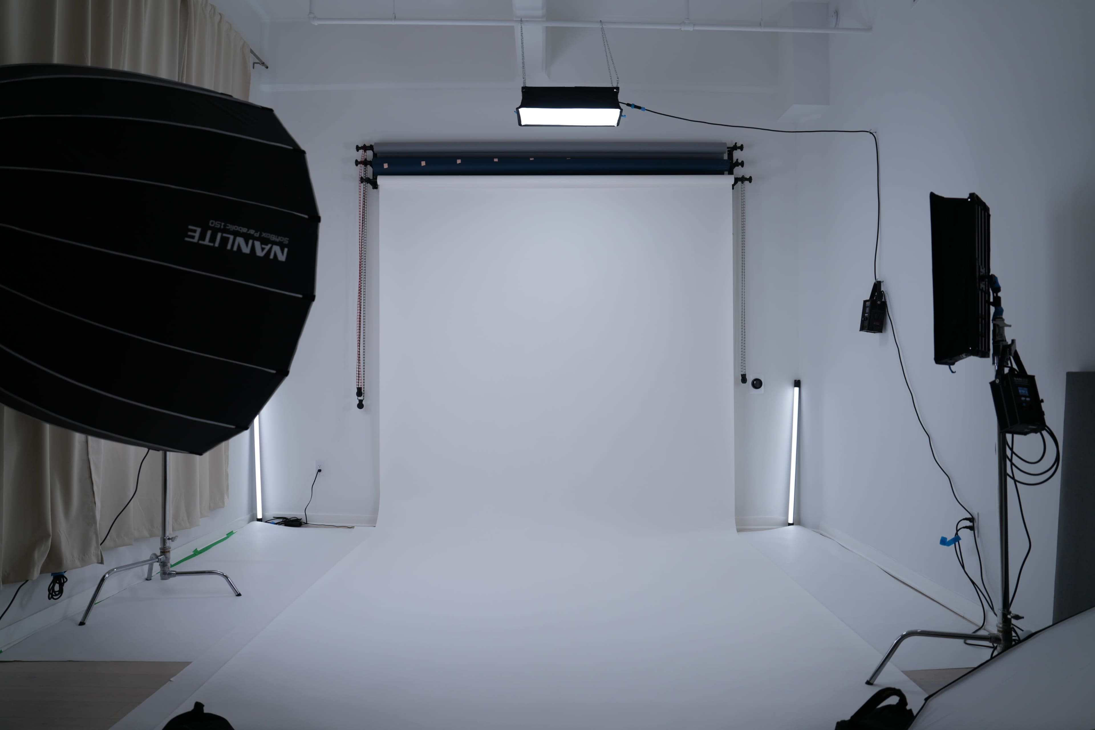 The image shows a photography studio with a white backdrop, softbox lighting, and various equipment along the walls.