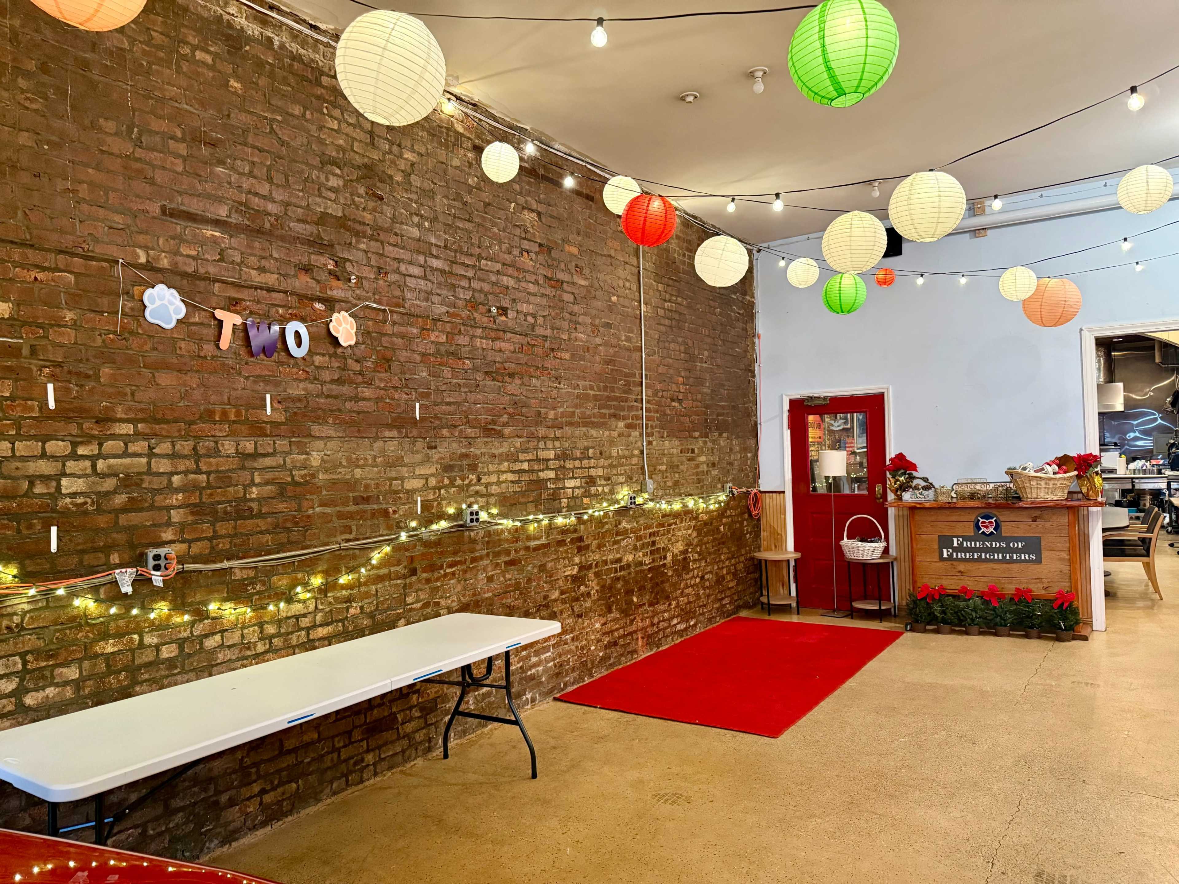The image shows a room with a brick wall, a red carpet leading to a counter, and colorful paper lanterns hanging from the ceiling, along with fairy lights and a table on one side.