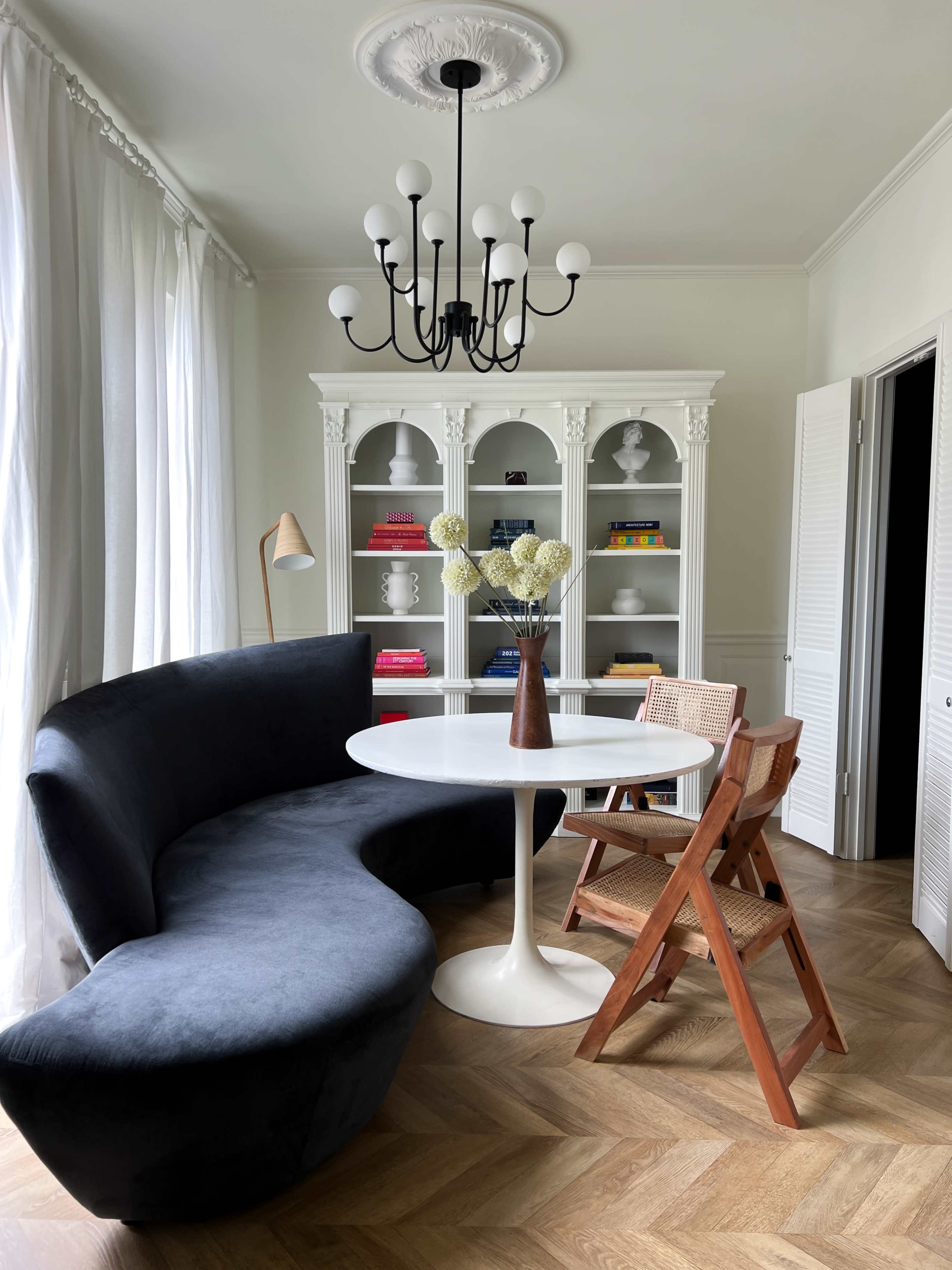The image shows a modern living space with a curved dark sofa, a round white dining table, and a pair of wooden chairs next to a white bookshelf filled with colorful books.