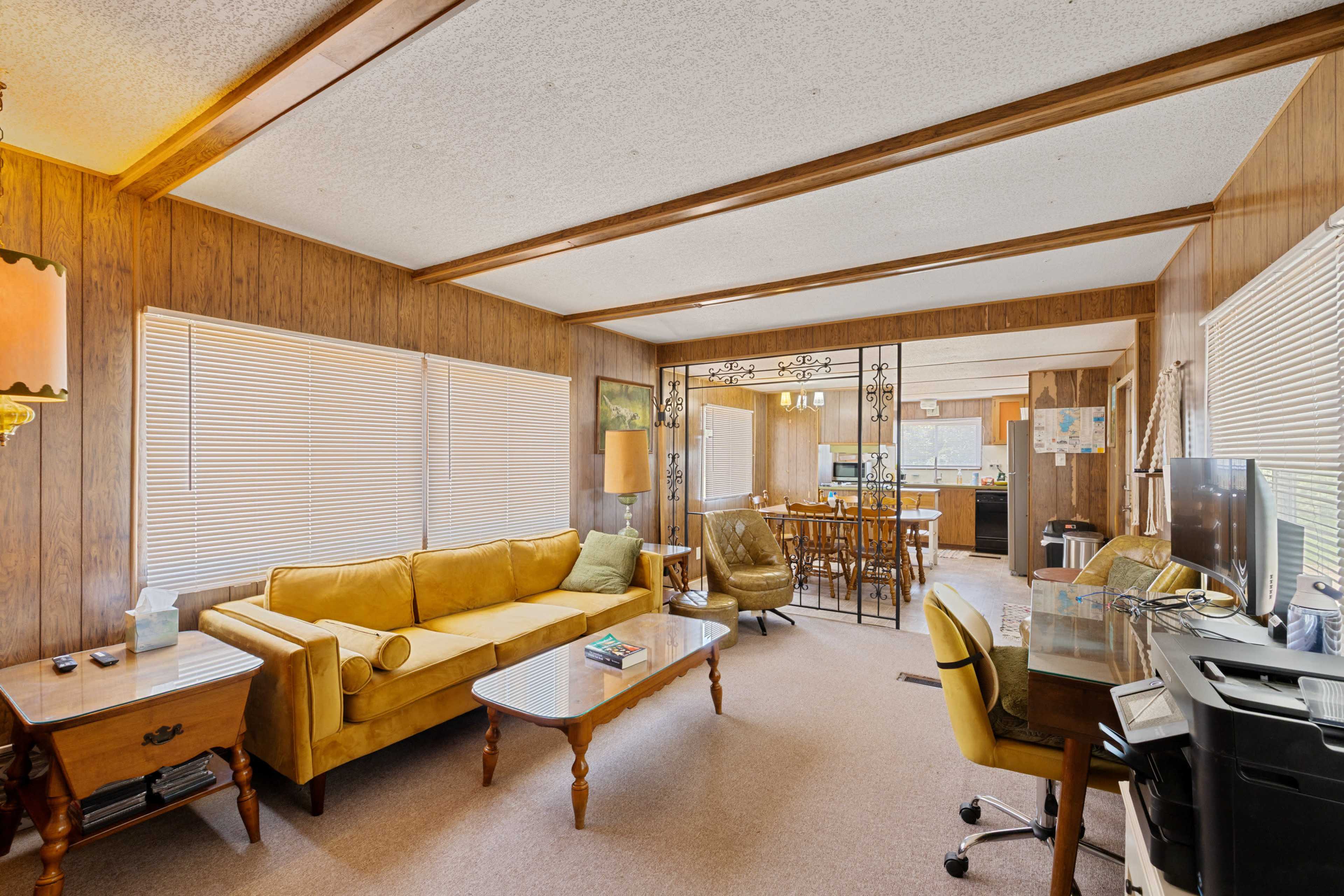 The image shows a living room with a yellow sofa, wooden paneling on the walls, and a visible dining area and kitchen in the background.