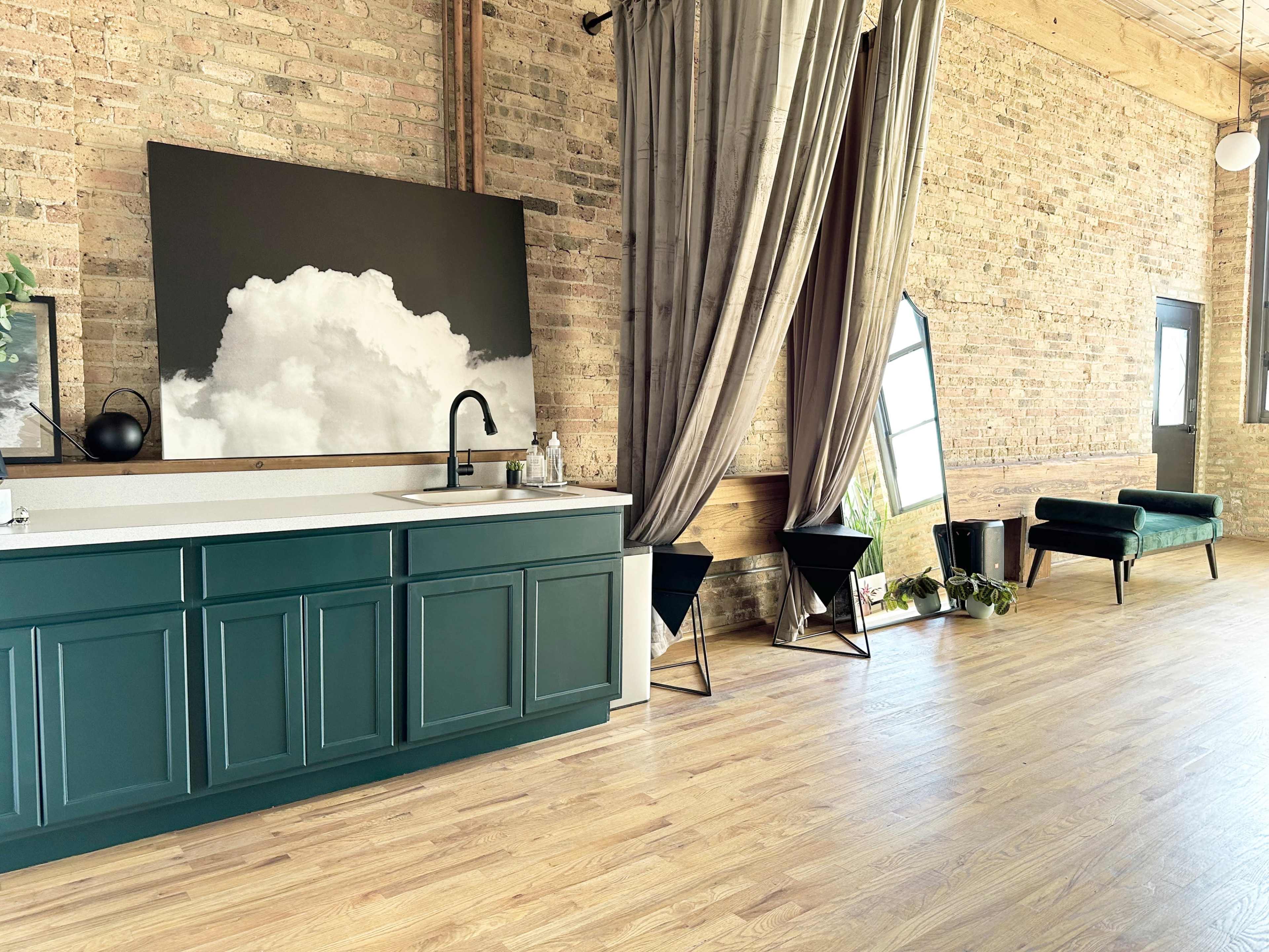 The image shows a modern interior featuring a green kitchen cabinet, a white countertop, a large black and white cloud painting, and a mix of decorative elements including curtains and furniture against a brick wall.