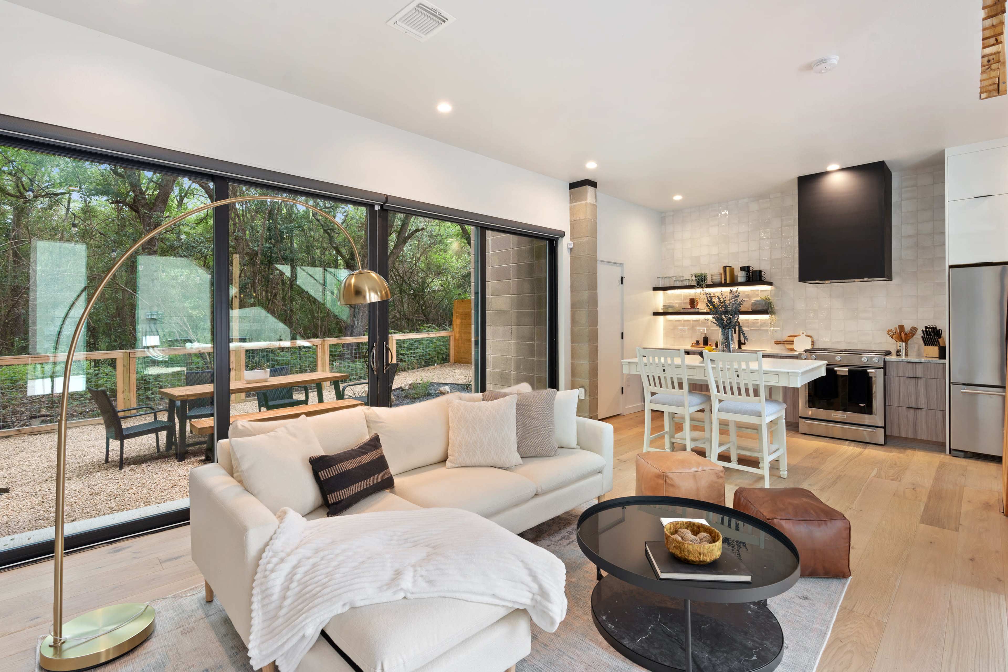 A modern living space with a white sofa, a circular coffee table, and a kitchen area featuring stainless steel appliances and bar stools, all overlooking a patio with a view of trees.