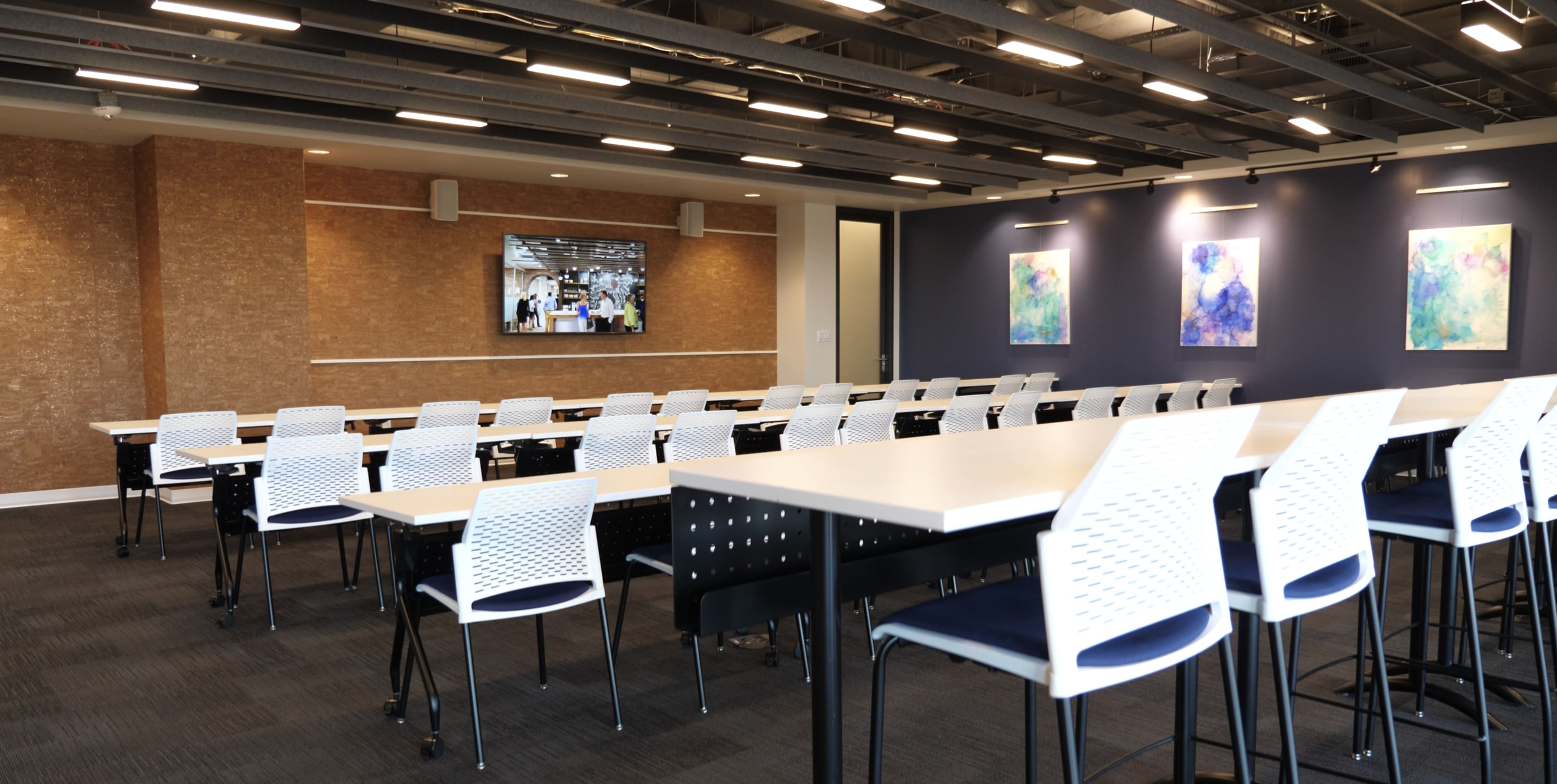 A modern conference room with long tables, white chairs, and a large TV screen displaying a presentation on the wall.