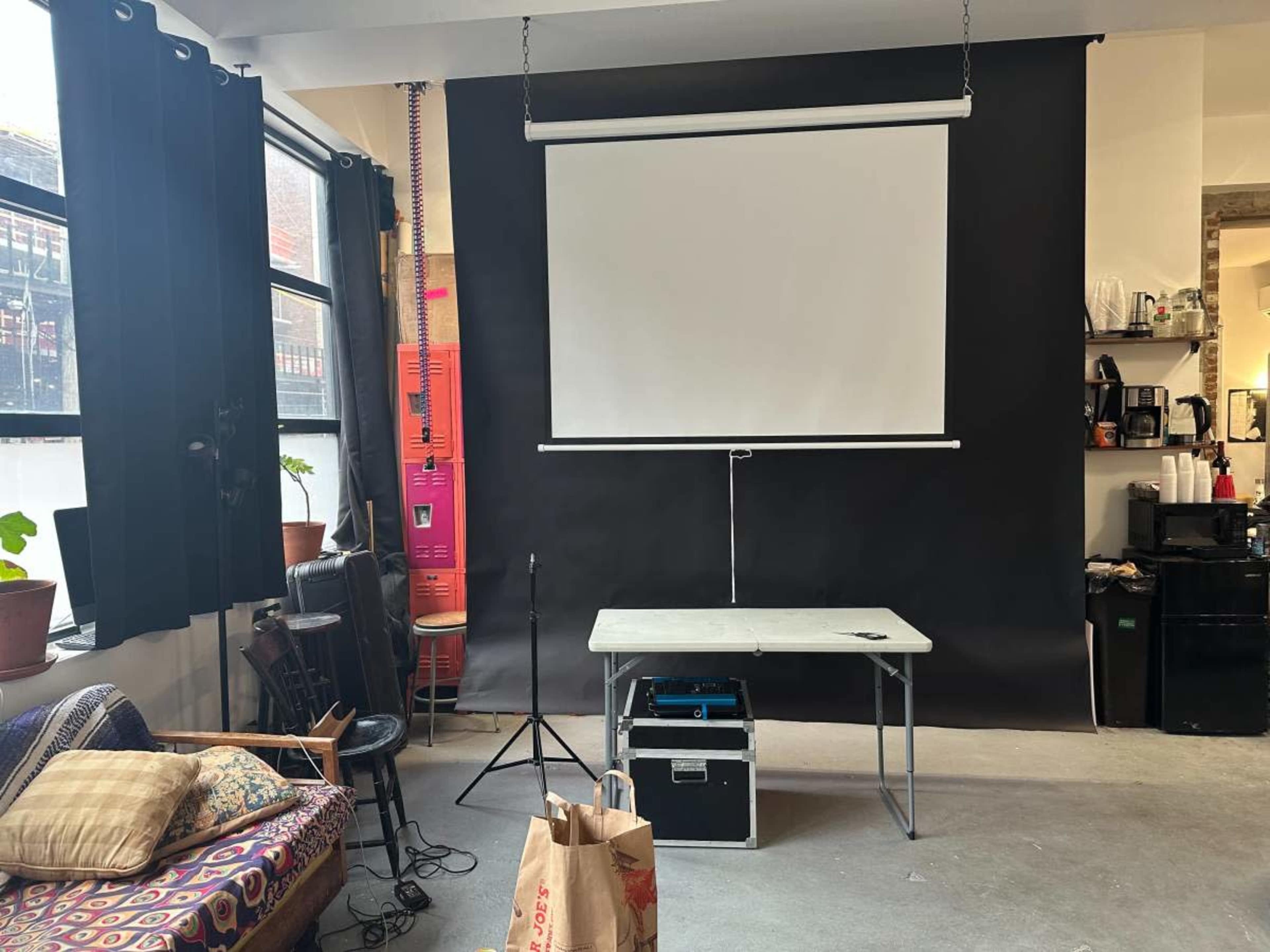 The image shows a room with a black backdrop, a white projection screen, a table, a microphone stand, and various furniture items including a couch and a kitchen area.