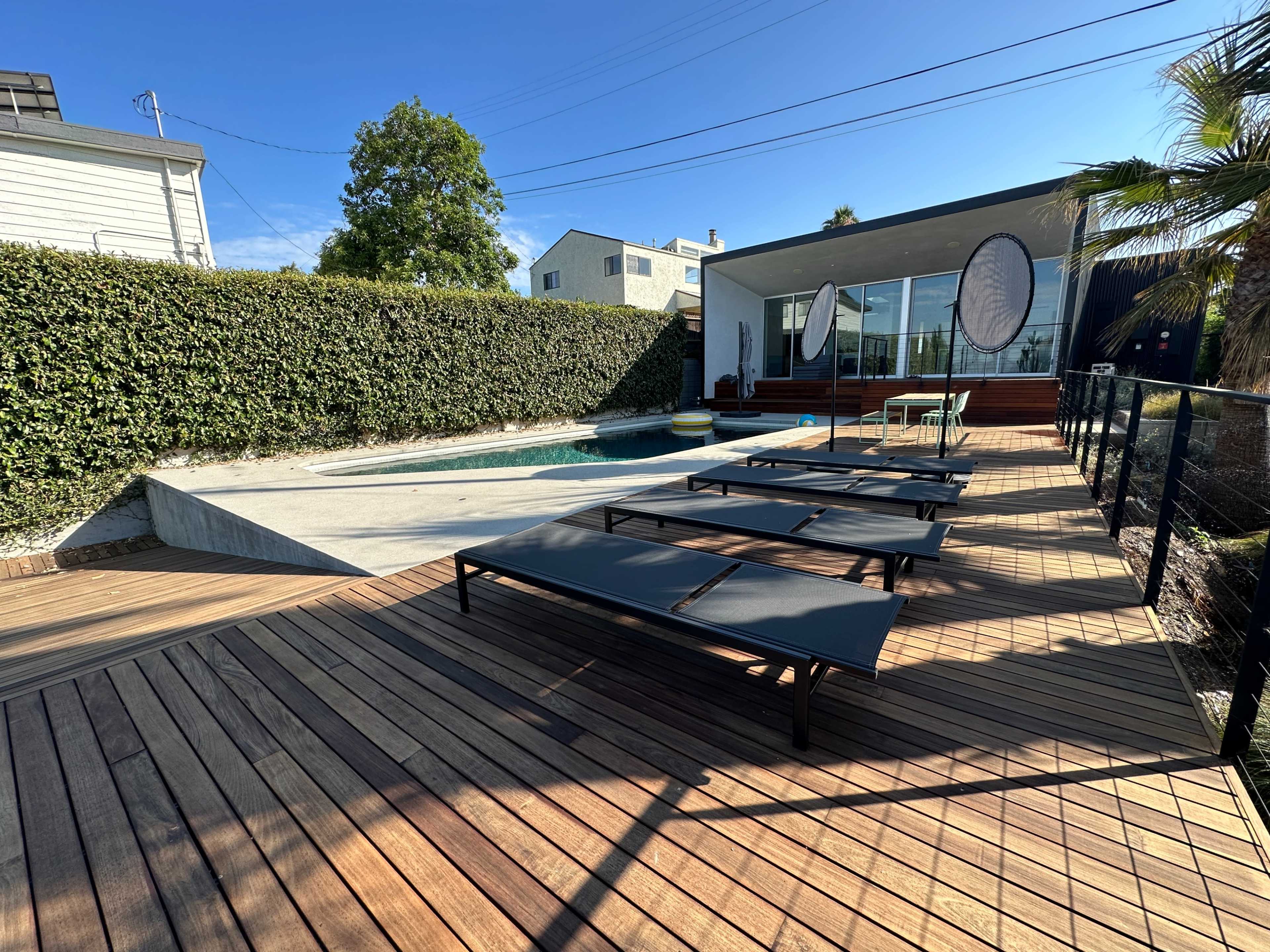 A modern pool area with wooden decking, lounge chairs, and a backdrop of lush greenery and a contemporary house.