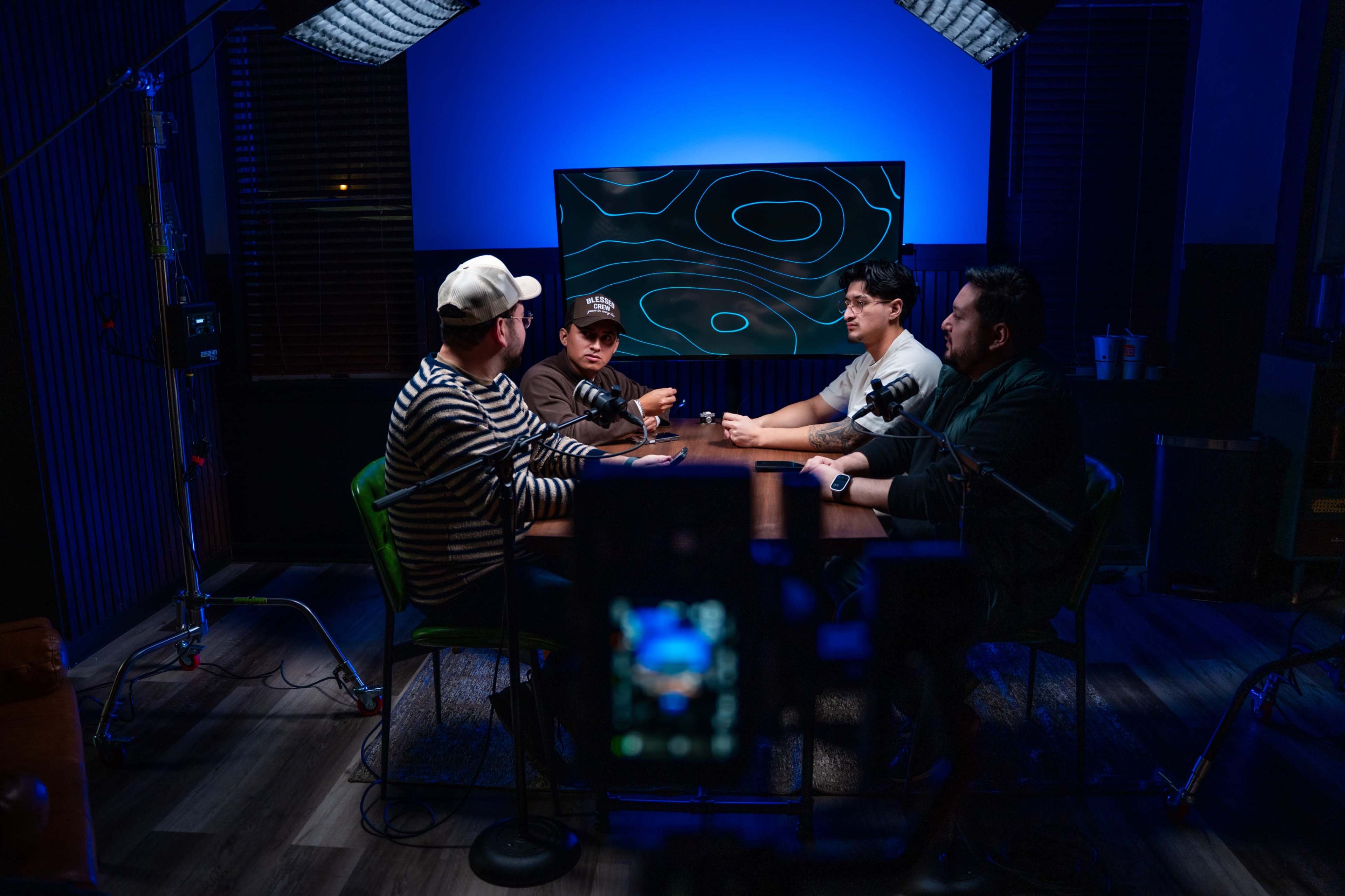 Four individuals sit around a table in a well-lit room, with a large screen displaying a map-like design in the background.