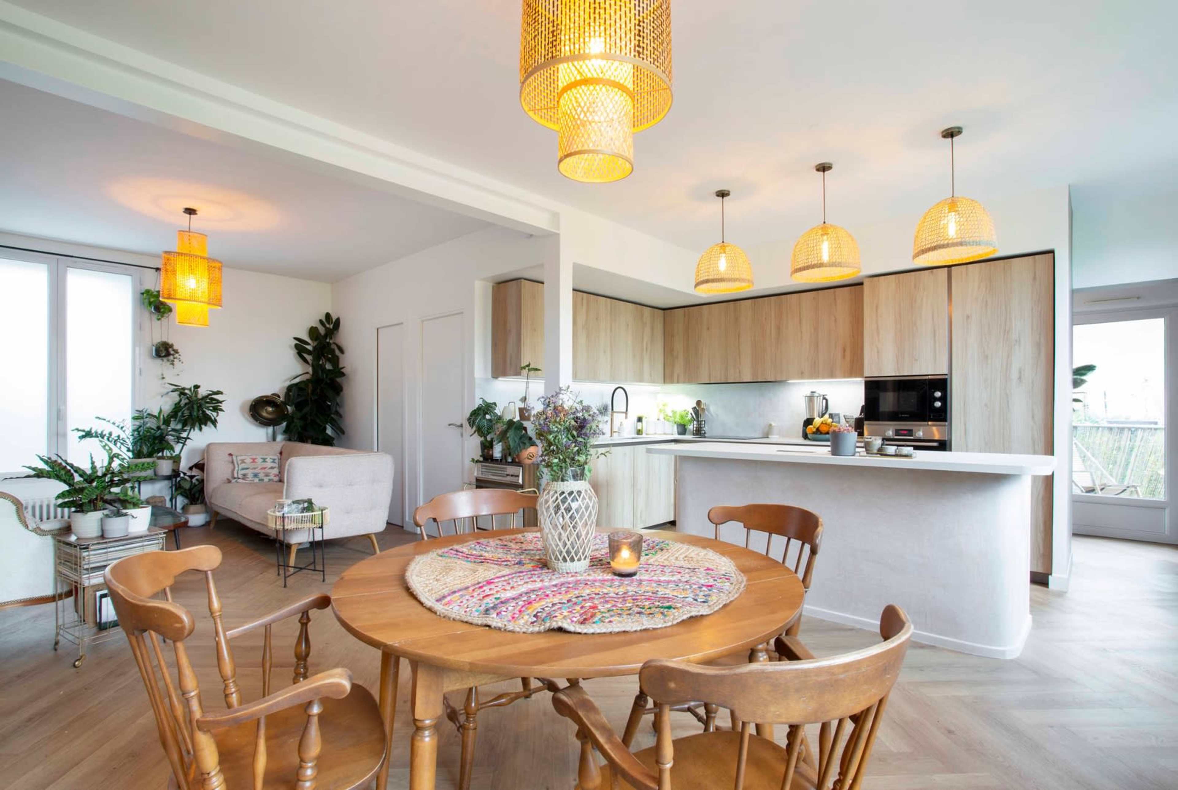 A modern dining area features a round wooden table surrounded by chairs, with a kitchen and plants visible in the background.