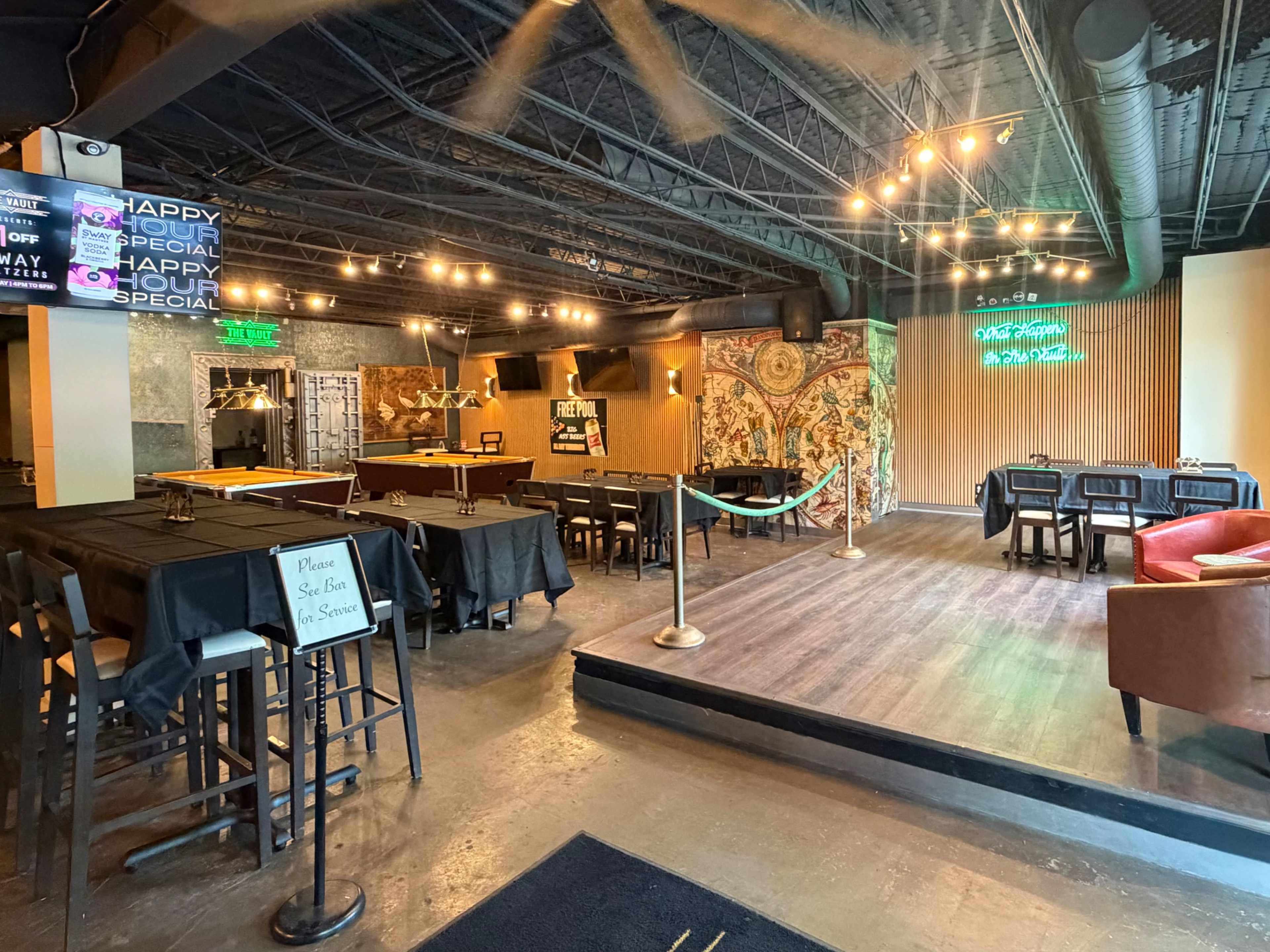 The interior of a bar is shown with dining tables, a small stage area, and a bar counter in the background, all under decorative lighting.