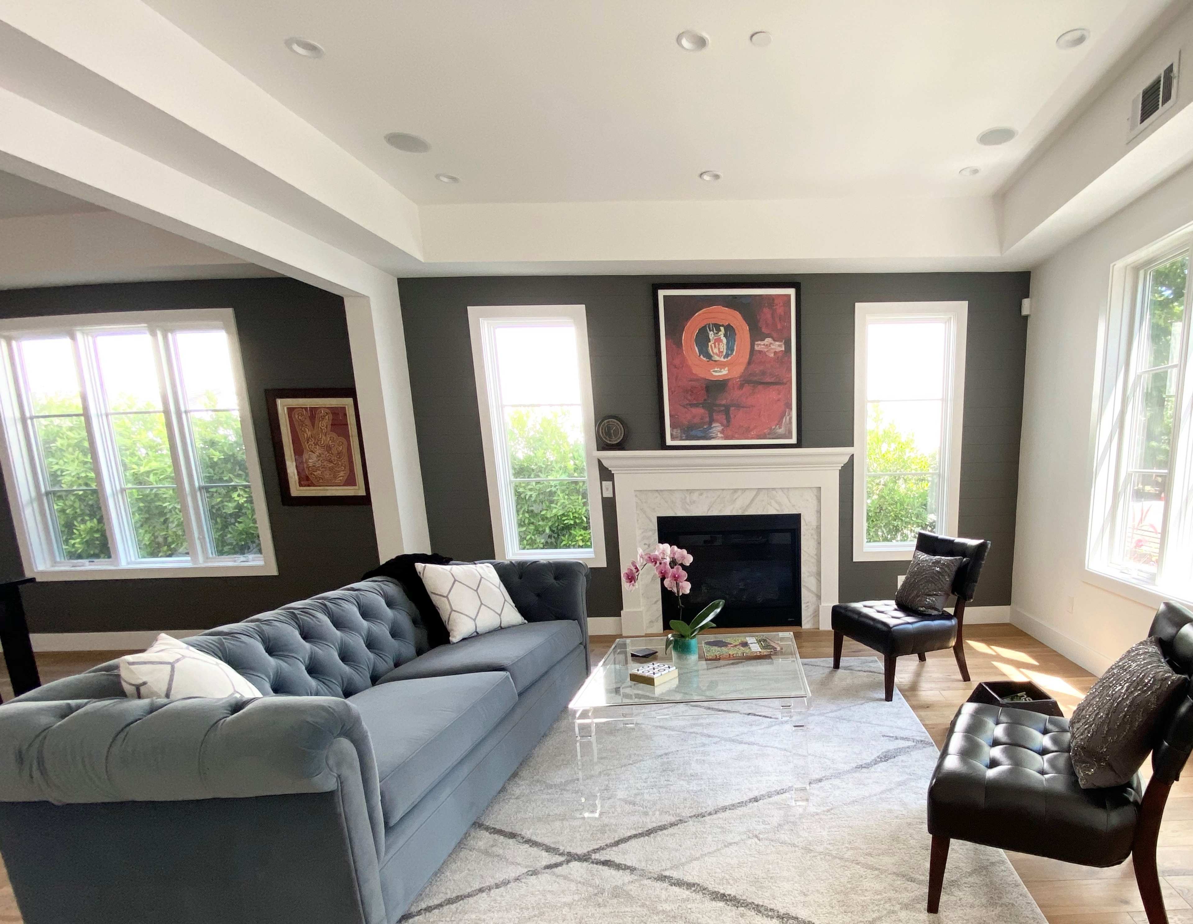 A modern living room features a blue tufted sofa, a glass coffee table, a fireplace, and large windows with green foliage outside.