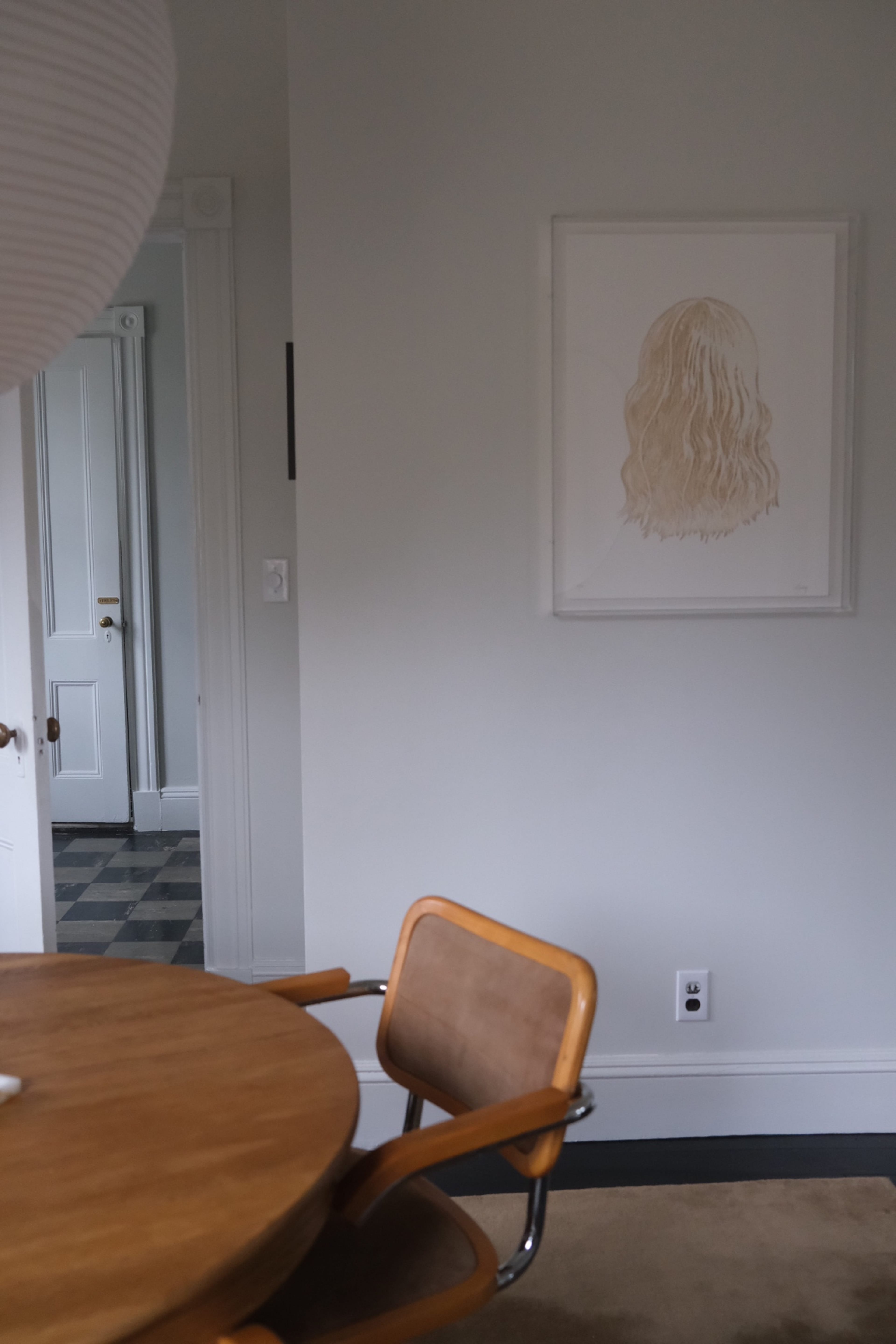 A wooden dining table is positioned in a room with a gray wall, featuring a framed artwork of a hair illustration and a doorway leading to another space.
