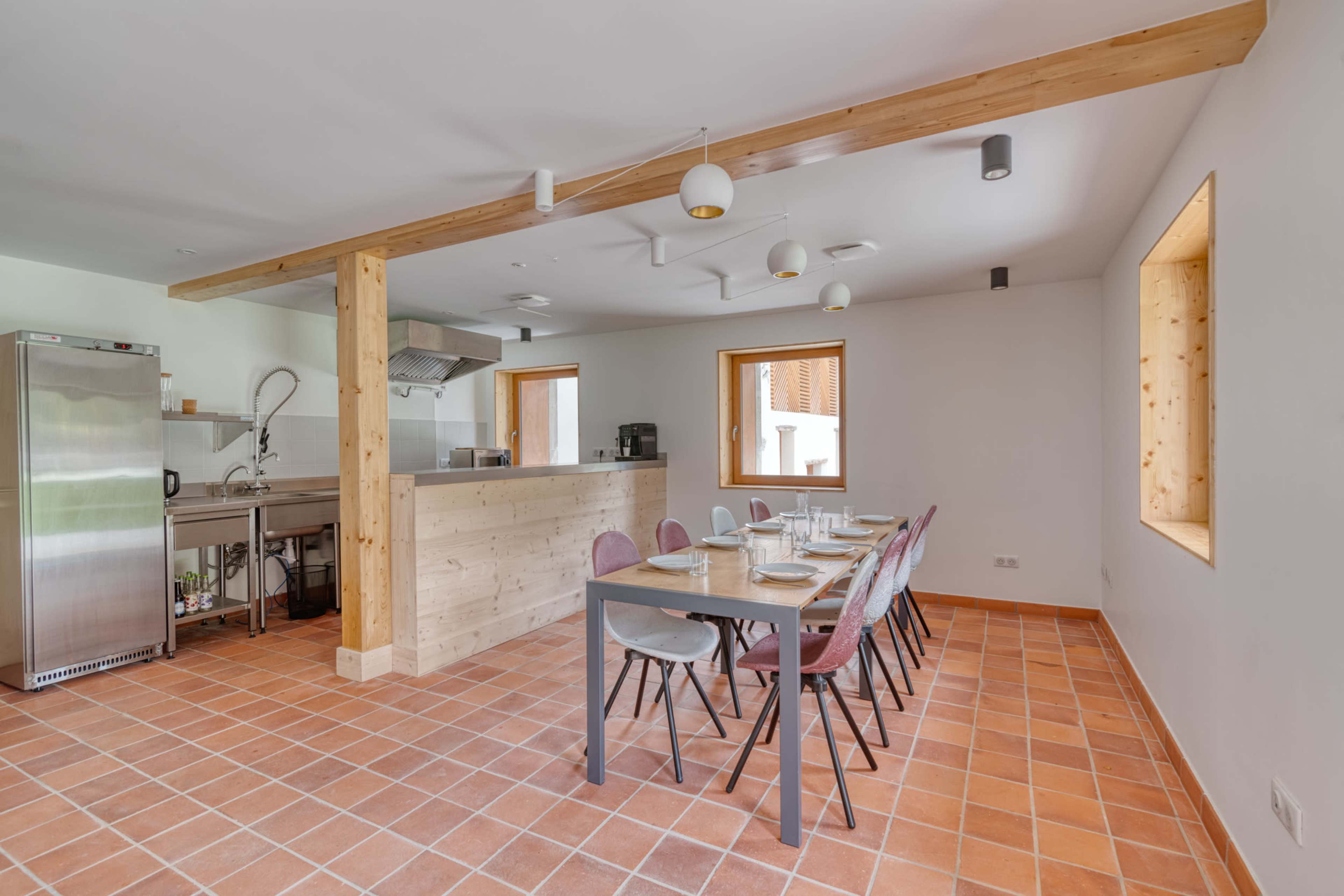 The image shows a modern, minimalist kitchen and dining area with a long table set for a meal, light wood accents, and tiled flooring.