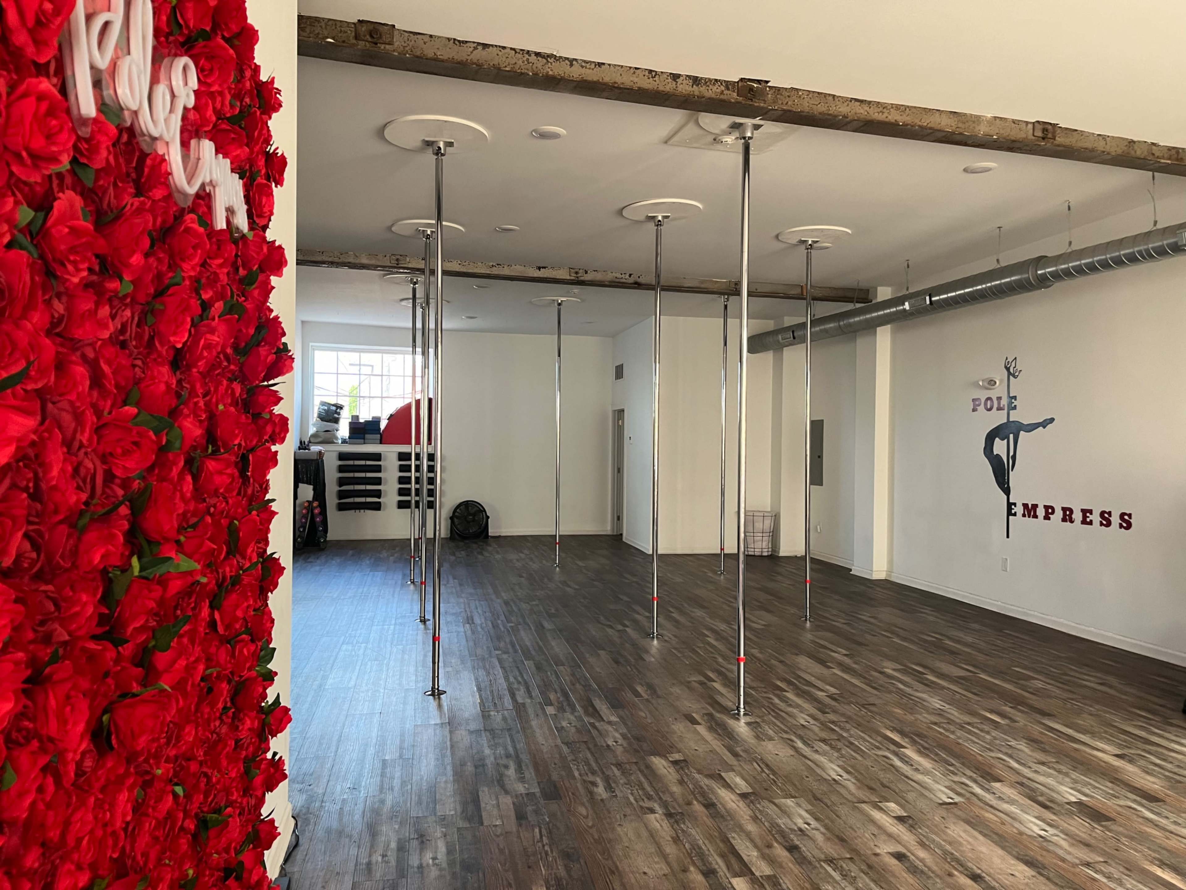 The image shows an interior of a dance studio with wooden floors, several dance poles, and a decorative wall filled with red roses.